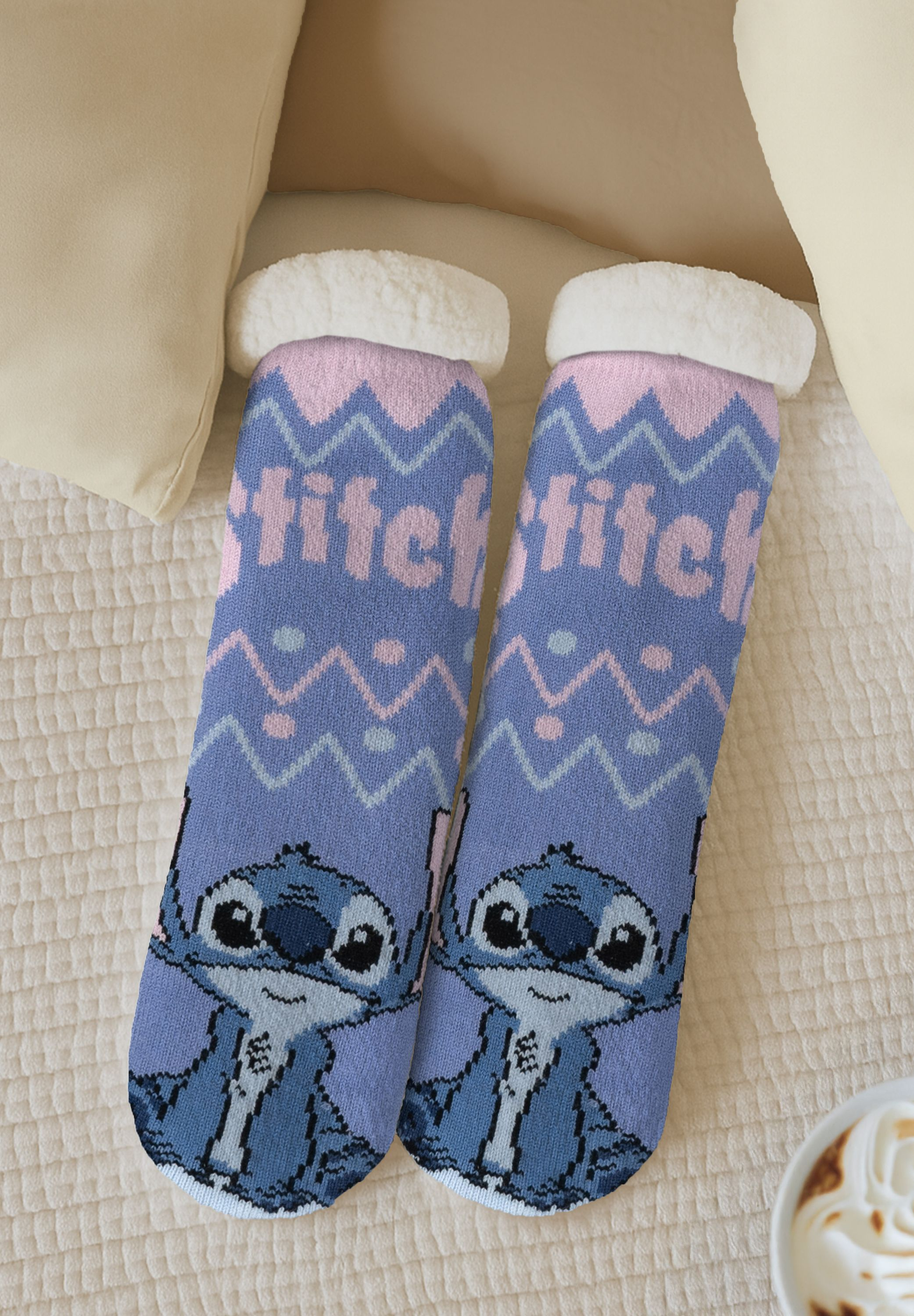 Zwei blaue Socken mit Stitch-Charakter-Design, weiße flauschige Oberteile, auf einem weißen Bett, mit neutralen Kissen und einer Kaffeetasse.