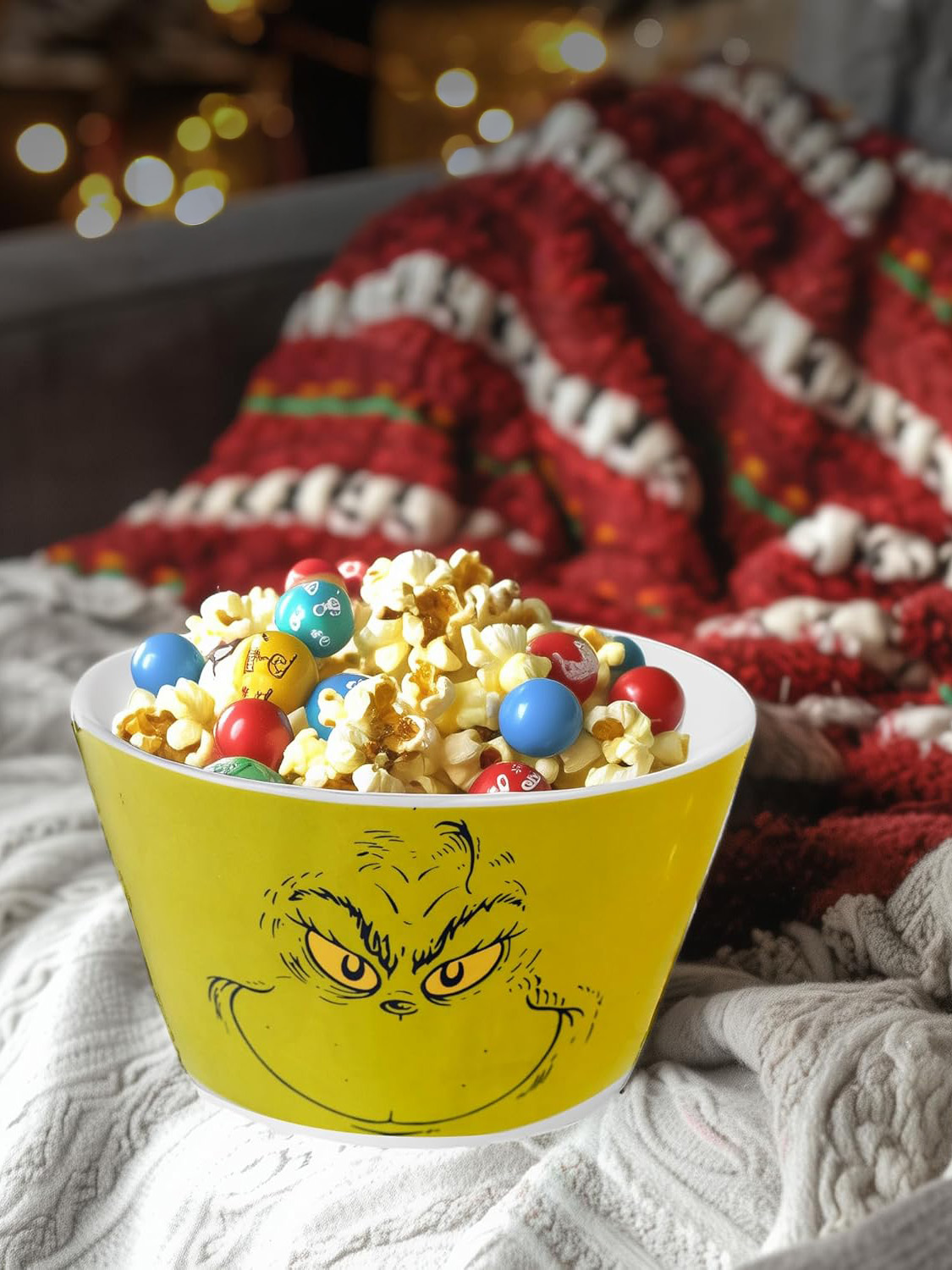 Eine gelbe Grinch-Schüssel mit Popcorn und Süßigkeiten. Im Hintergrund eine rot-weiß karierte Decke mit verschwommenen Lichtern.