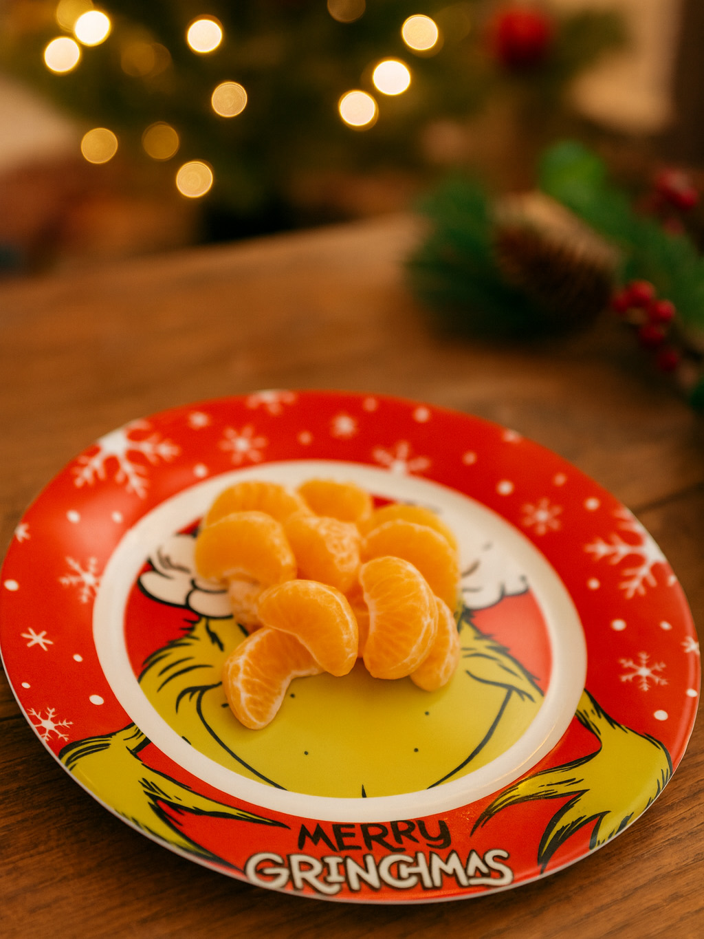 Ein Teller mit Mandarinensegmenten. Roter Rand mit Schneeflocken. Der Teller hat ein Grinch-Design. Lichter des Weihnachtsbaums im Hintergrund.