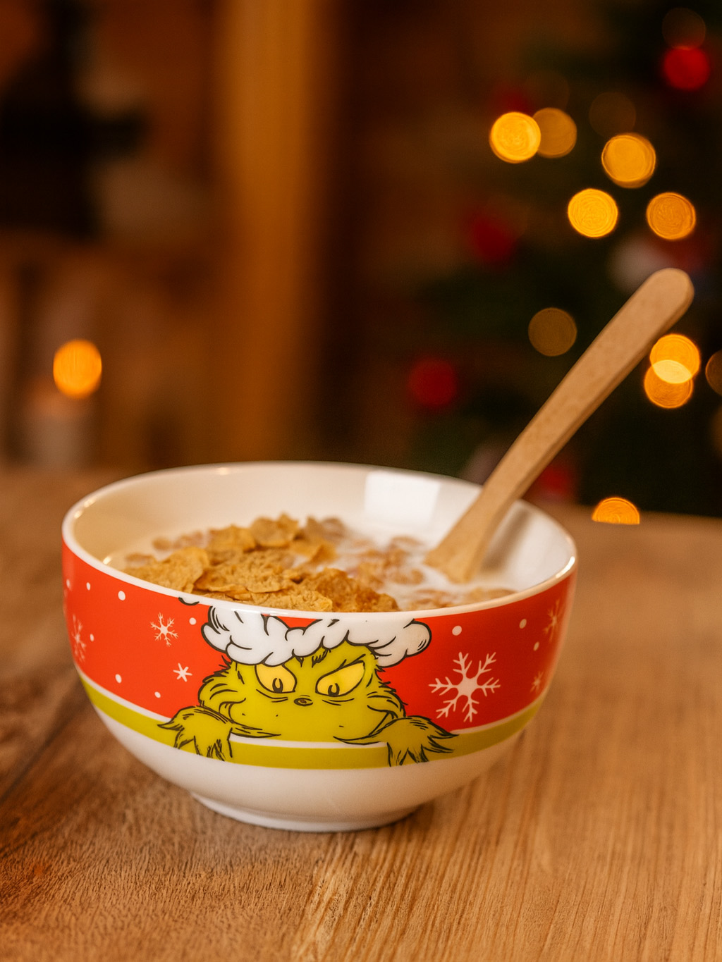 Eine Schüssel Müsli mit dem Grinch. Die Schüssel steht auf einem Holztisch, mit einem Holzlöffel darin. Im Hintergrund sind Weihnachtslichter zu sehen.