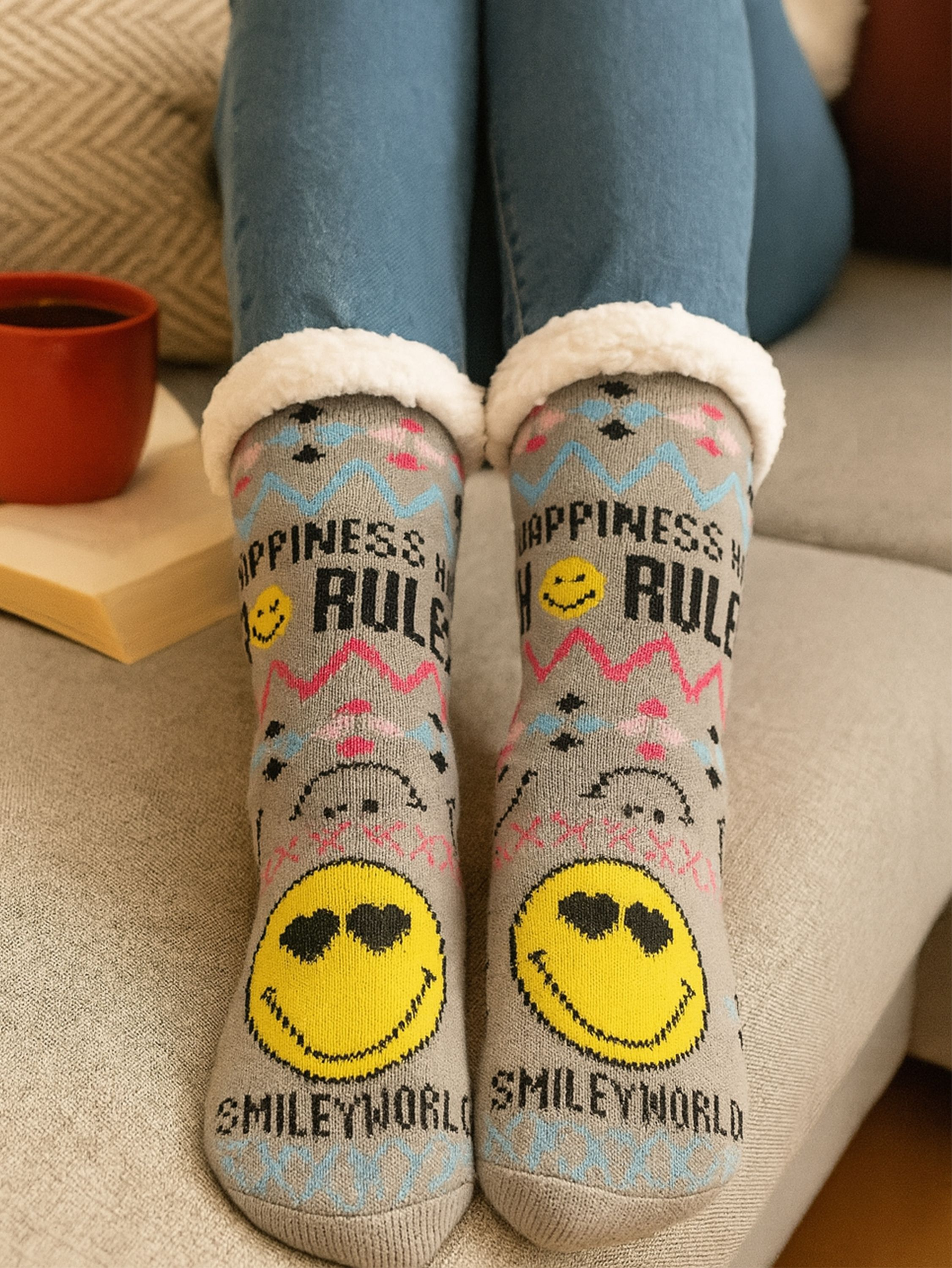 Person trägt blaue Jeans und graue Socken mit Smiley-Gesichtern. Auf der linken Seite sind eine rote Tasse und ein Buch zu sehen.
