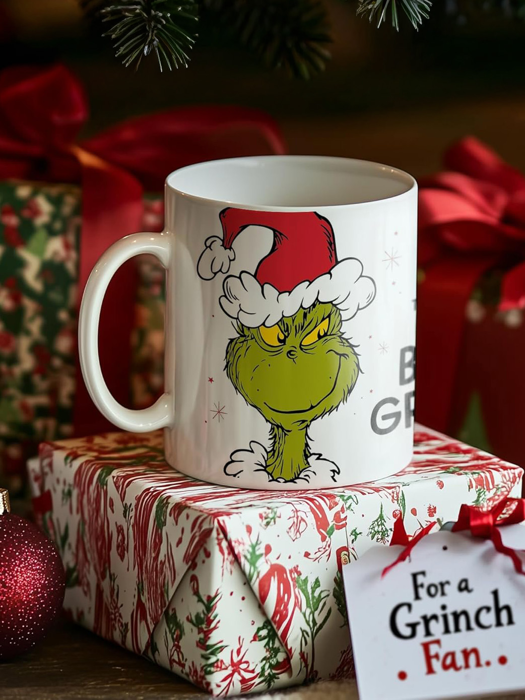 Eine Tasse mit Grinch-Design steht auf einer Geschenkbox. Andere verpackte Geschenke und Ornamente sind sichtbar, mit einem Etikett 'Für einen Grinch-Fan'.