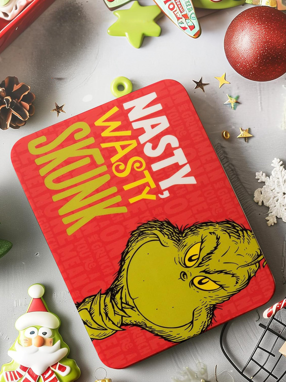 Eine rote Geschenkbox zum Thema Grinch mit den Worten 'Nasty, Wasty, Skunk'. Weihnachtsornamente und Kekse sind verstreut.