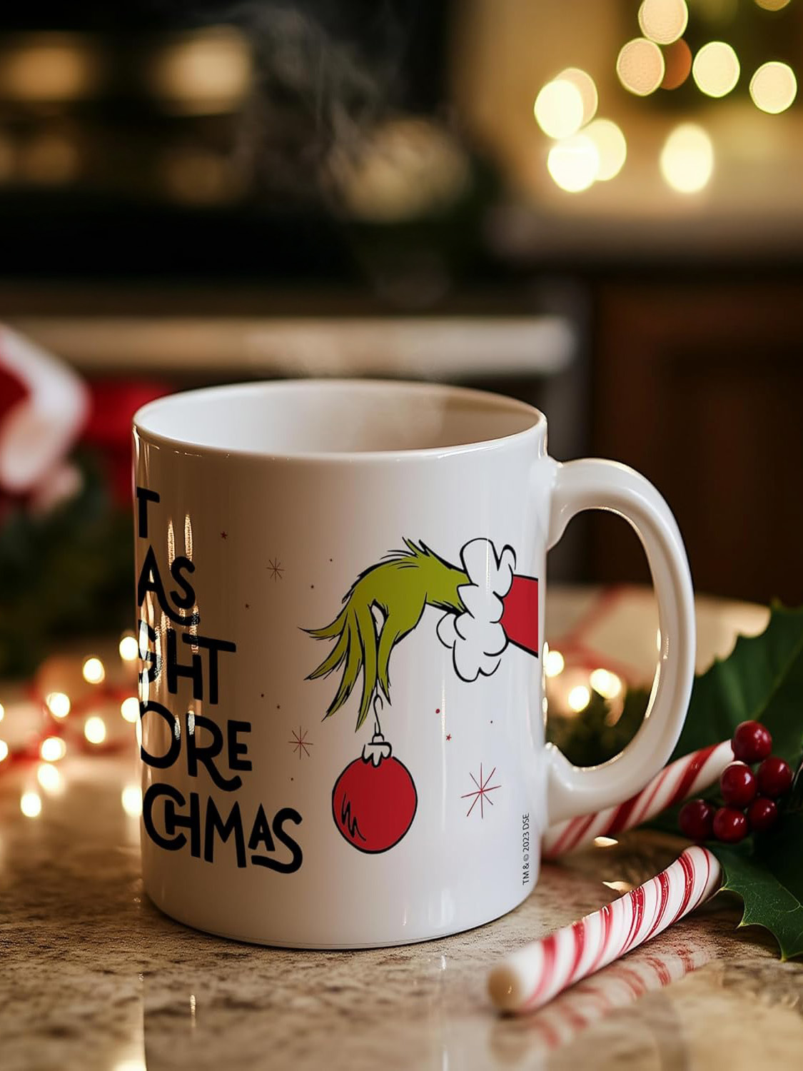 Weiße Tasse mit Grinch-Design, Dampf steigt auf. Zuckerstangen und Stechpalmen liegen auf dem Tisch. Lichter im Hintergrund.