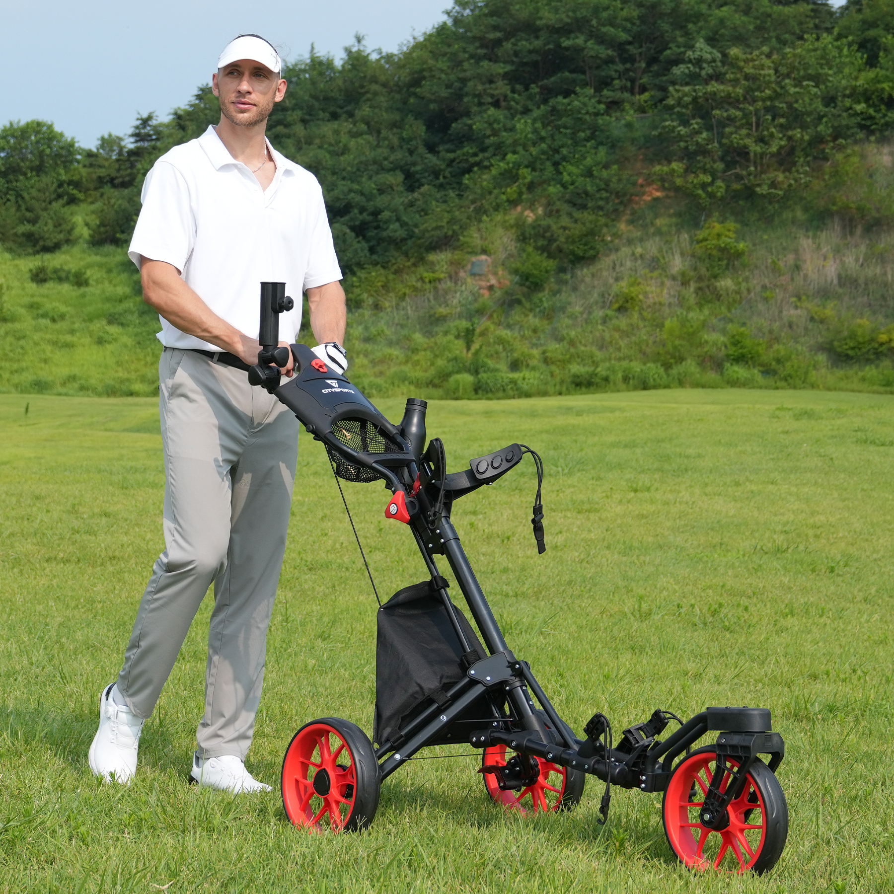 Mężczyzna w białej koszuli i daszku pcha wózek golfowy z czerwonymi kołami na zielonym polu golfowym.