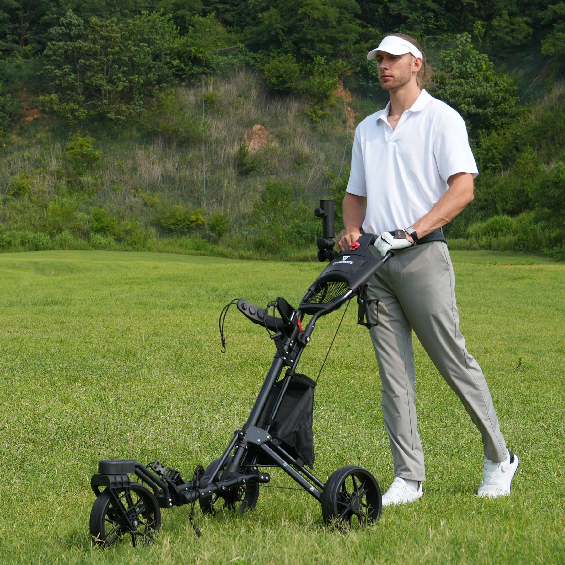Mężczyzna na polu golfowym pchający wózek golfowy. Ma na sobie białą koszulę, daszek i szare spodnie. Zielona trawa i drzewa w tle.