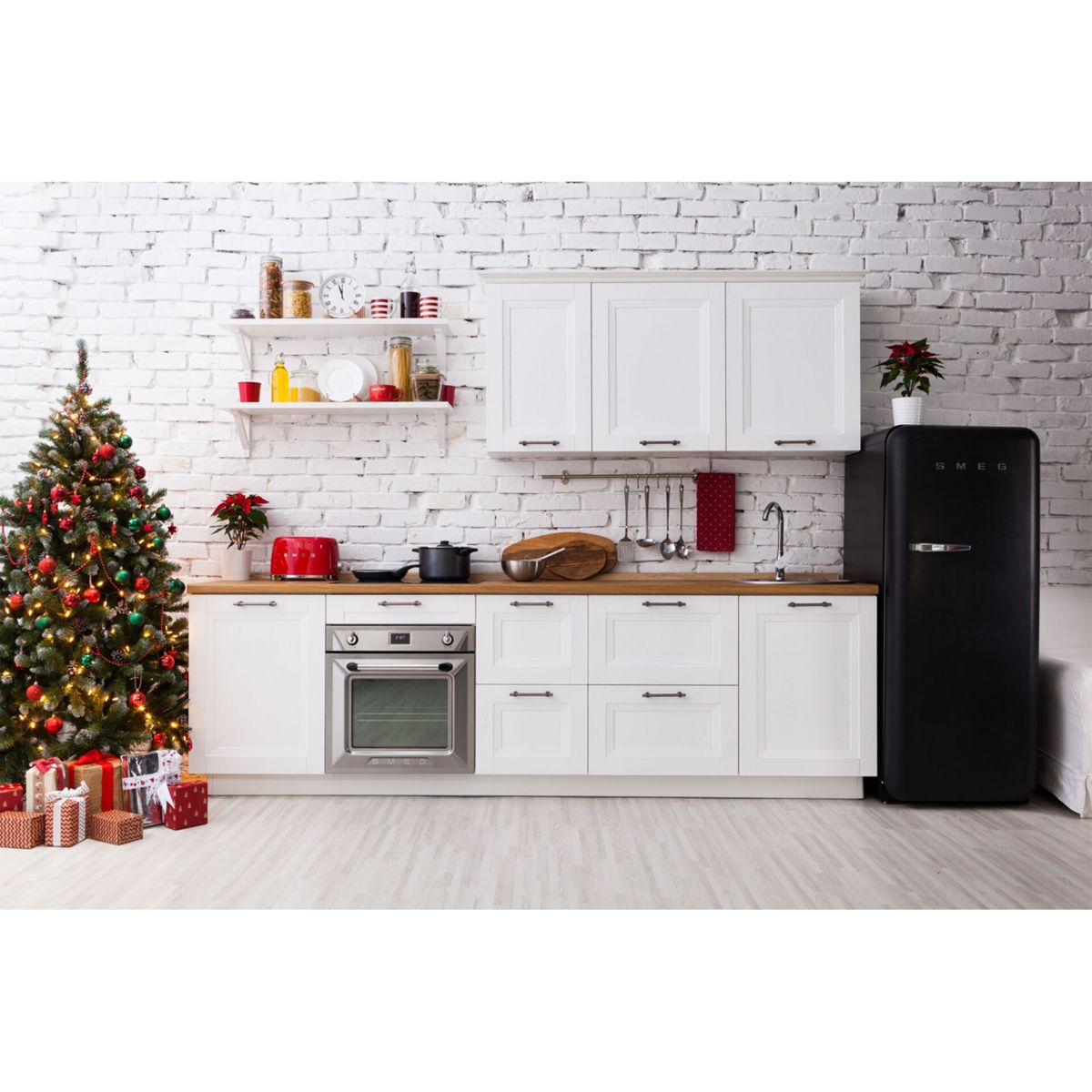 Kuchnia udekorowana na Boże Narodzenie z choinką, prezentami, białymi szafkami, czarną lodówką SMEG i czerwonym tosterem.