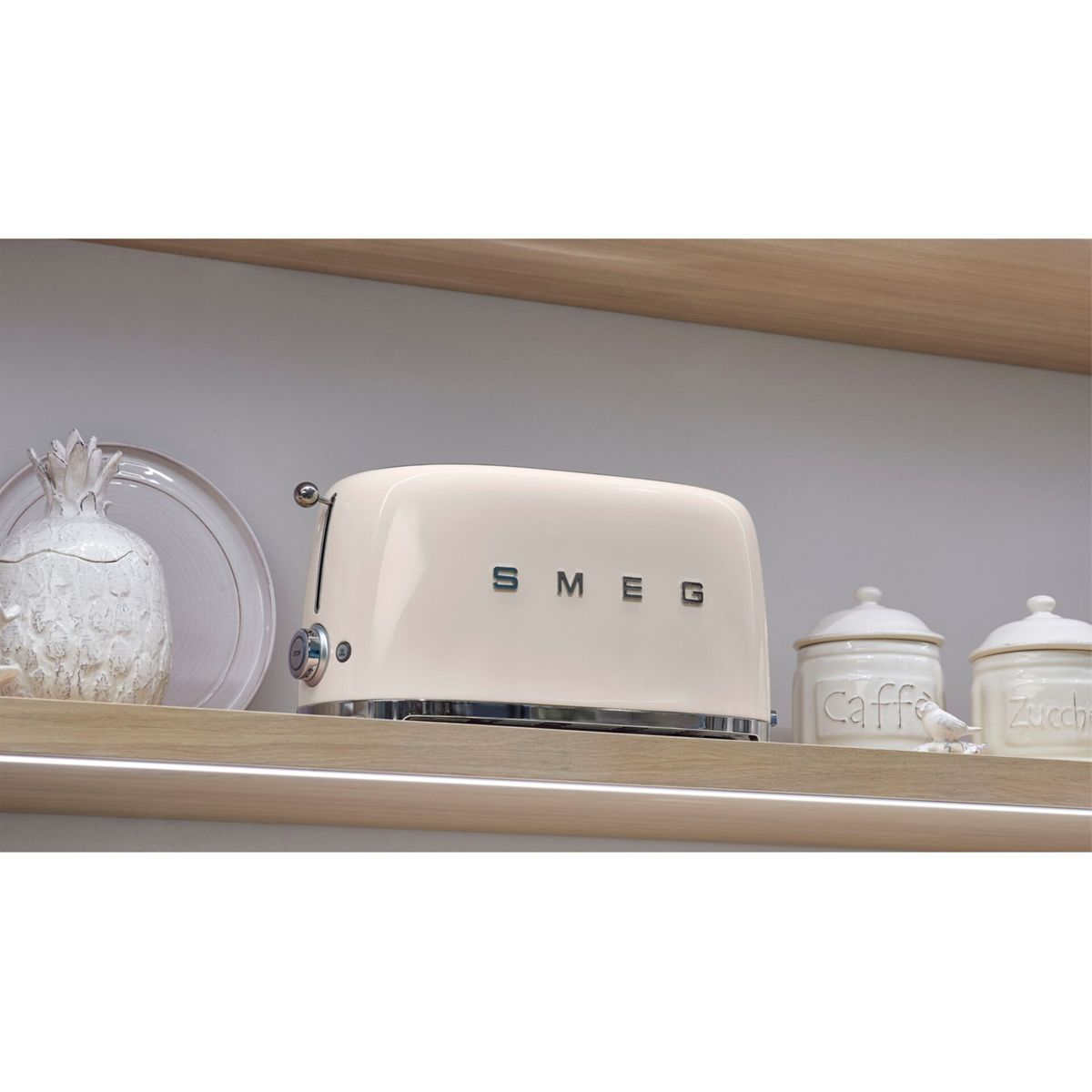 Kremowy toster Smeg na drewnianej półce z ananasowym słoikiem i dwoma pojemnikami z napisami 'Caffe' i 'Zuccher'.