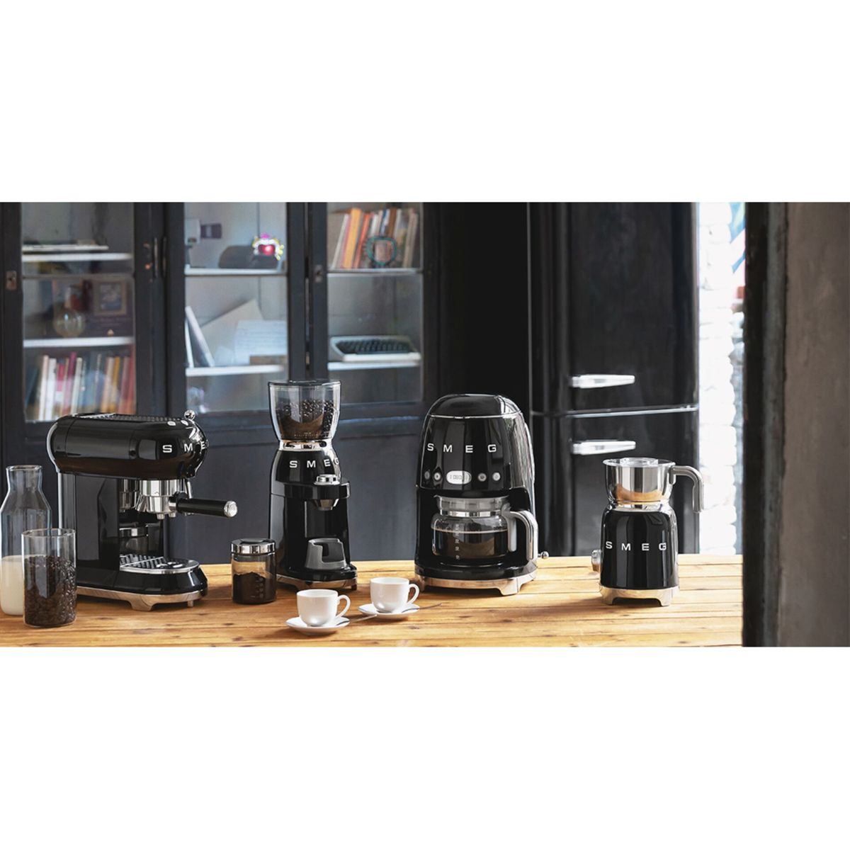 Kilka czarnych ekspresów do kawy SMEG na drewnianym stole, z filiżankami i szklaną butelką. Czarne szafki i lodówka w tle.