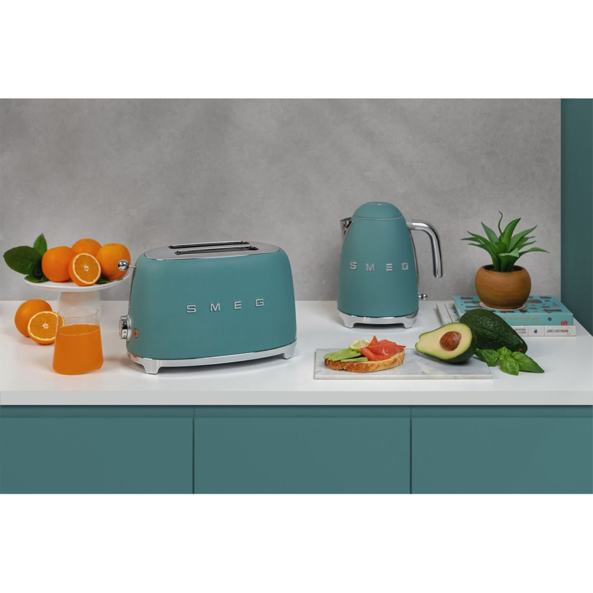 Turkusowy toster i czajnik SMEG stoją na białym blacie z pomarańczami, sokiem, tostami z awokado i rośliną doniczkową.