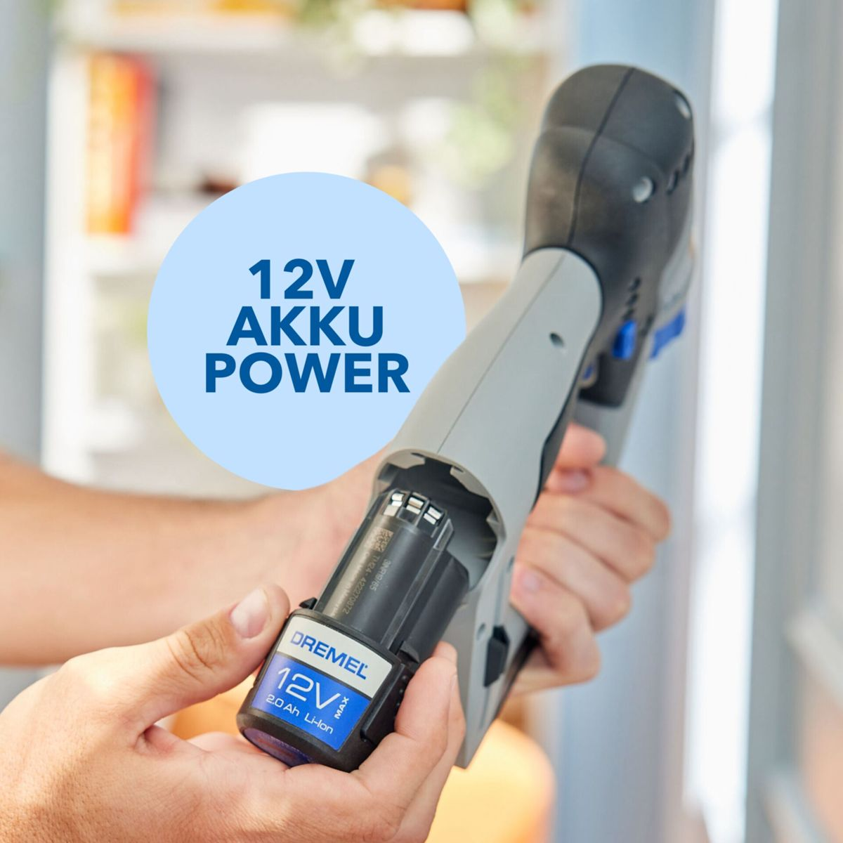 Main tenant un outil Dremel gris et noir, insérant une batterie 12V. Le texte sur l'image dit '12V AKKU POWER'.
