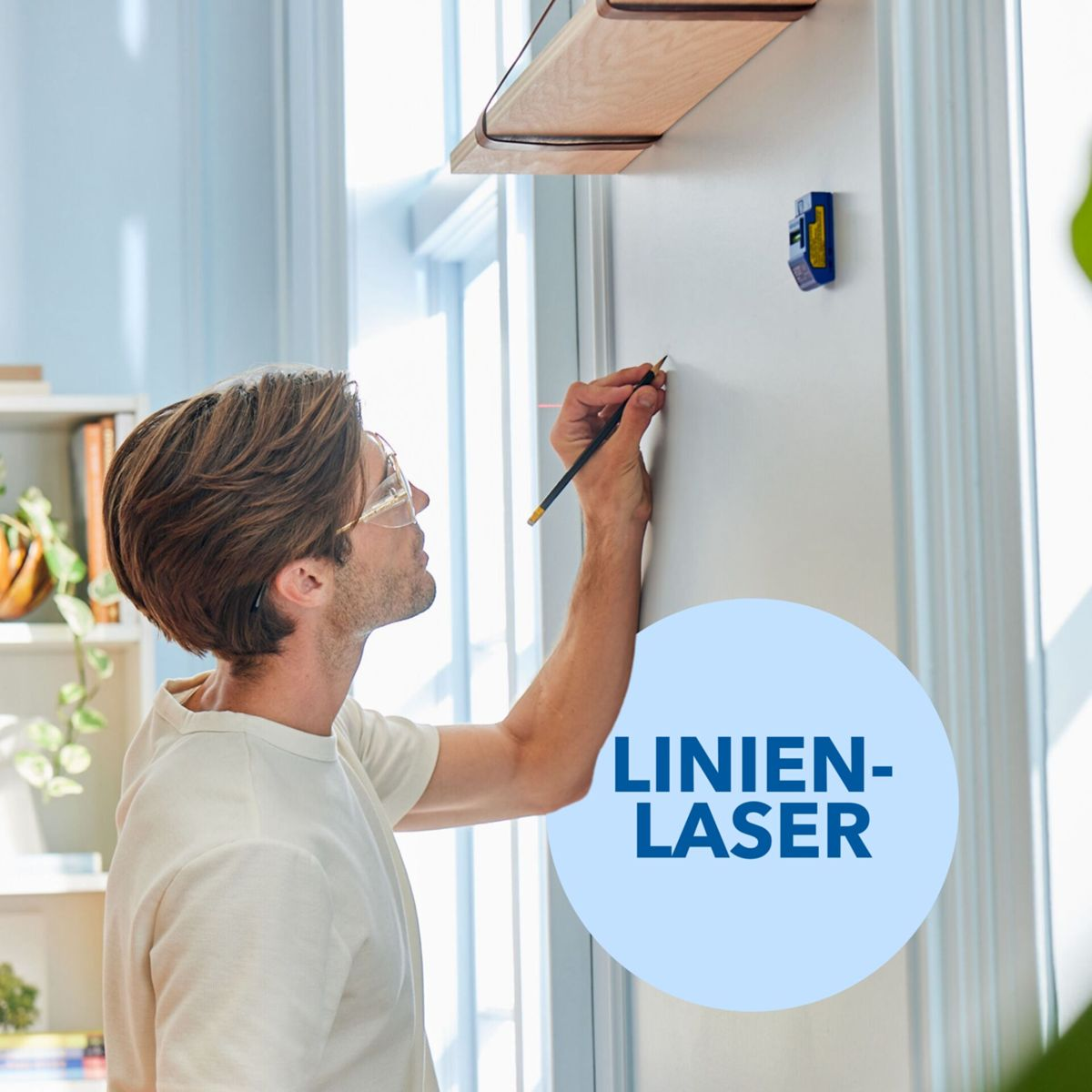 Un homme en lunettes dessine sur un mur blanc avec un crayon, près d'un niveau laser bleu et d'une étagère en bois.