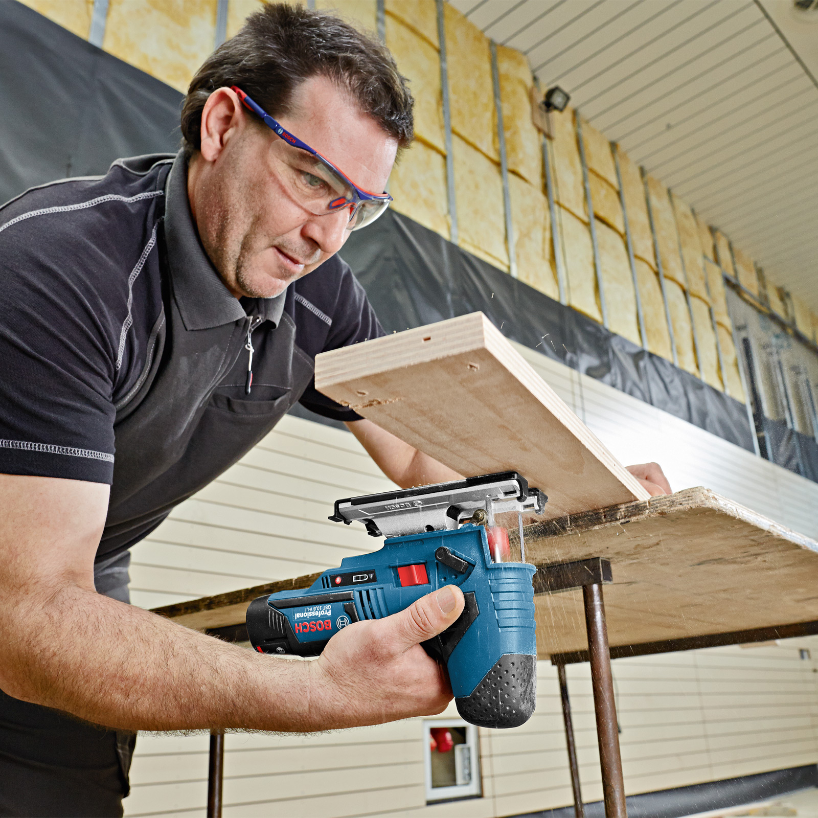 Ein Mann benutzt eine blaue Stichsäge, um eine Holzplatte zu schneiden. Er trägt eine Schutzbrille. Der Hintergrund ist eine Baustelle.