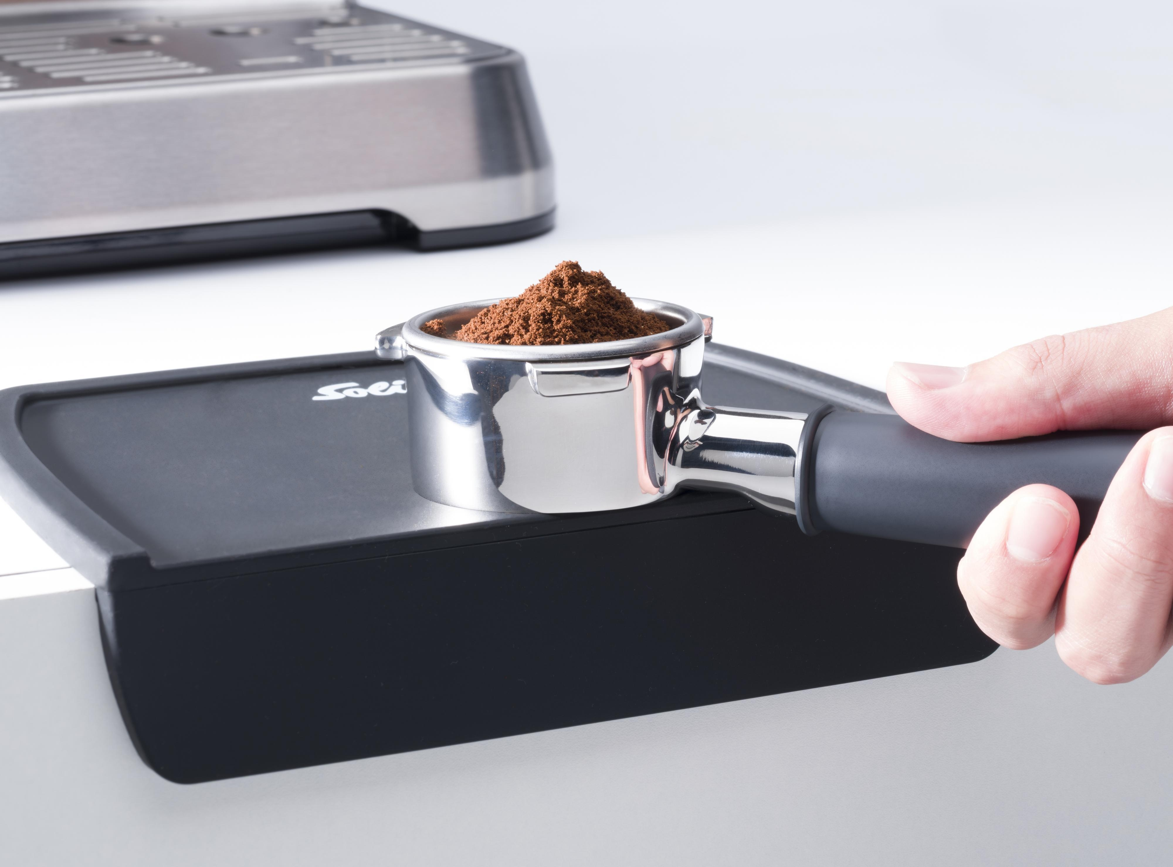 Eine Hand hält einen mit Kaffeepulver gefüllten Siebträger. Der Siebträger liegt auf einer schwarzen Oberfläche. Im Hintergrund befindet sich eine silberne Maschine.