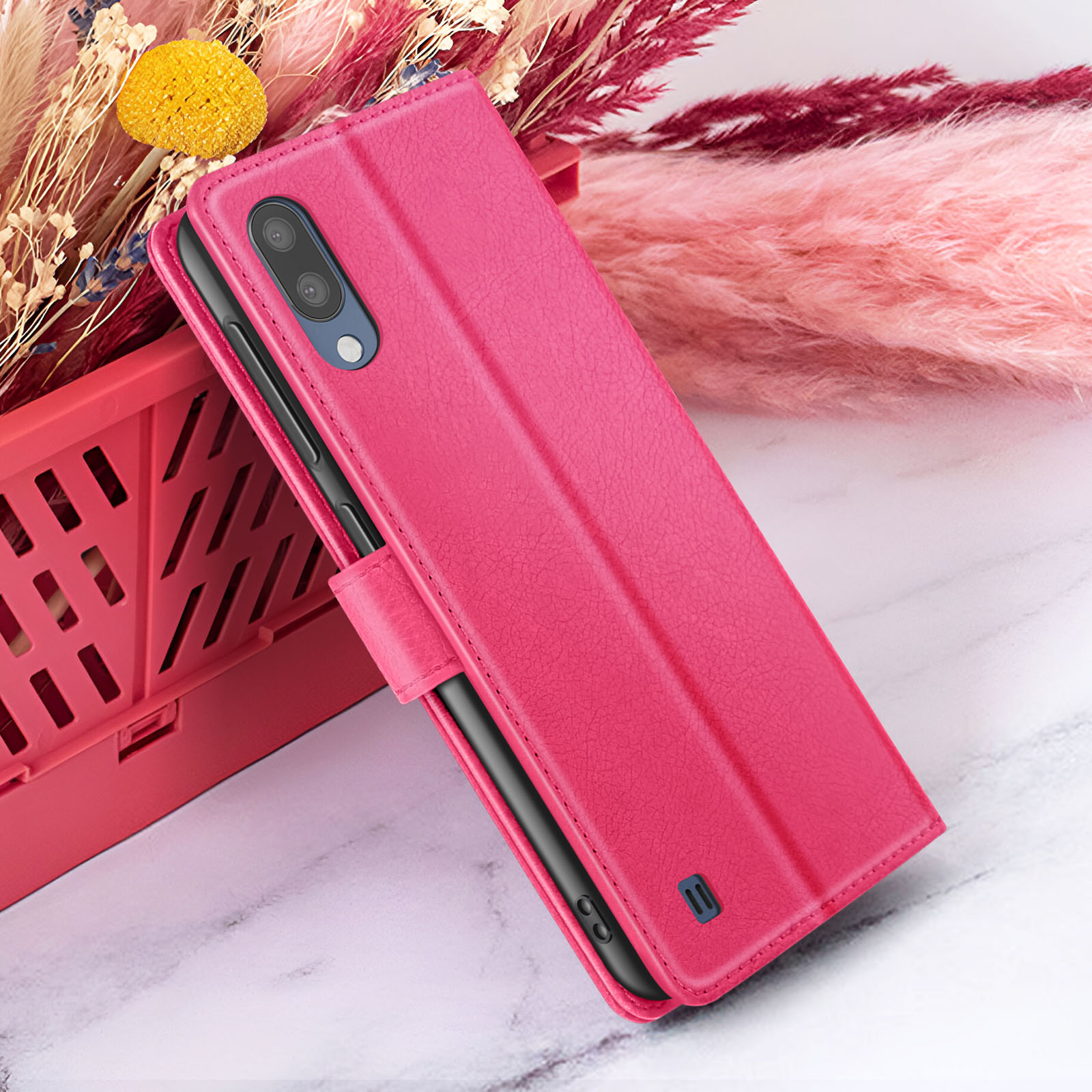Een magenta telefoonhoesje is open en onthult een zwarte telefoon. Het rust tegen roze bloemen en een roze mand.