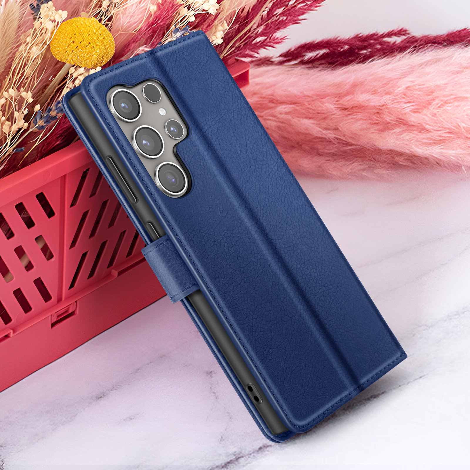 Een blauwe telefoonhoes met een telefoon erin ligt op een marmeren oppervlak. Bloemen en een rode mand op de achtergrond.