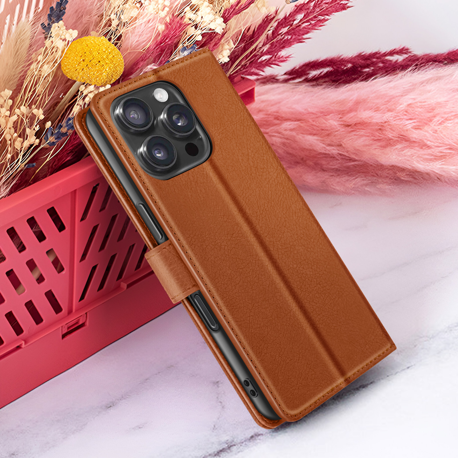 Un étui de téléphone en cuir marron avec une découpe pour l'appareil photo, posé sur un conteneur rouge, fond de fleurs séchées.