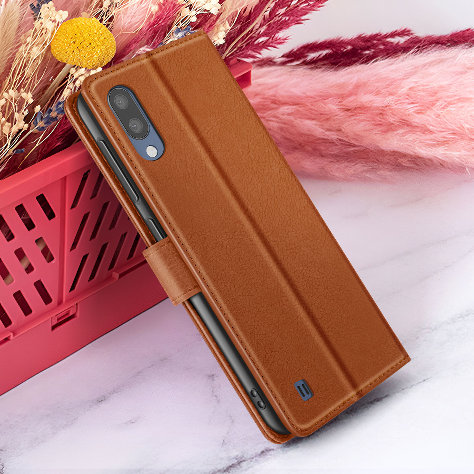Étui de téléphone en cuir marron sur une surface en marbre. Fleurs roses et panier rouge en arrière-plan.