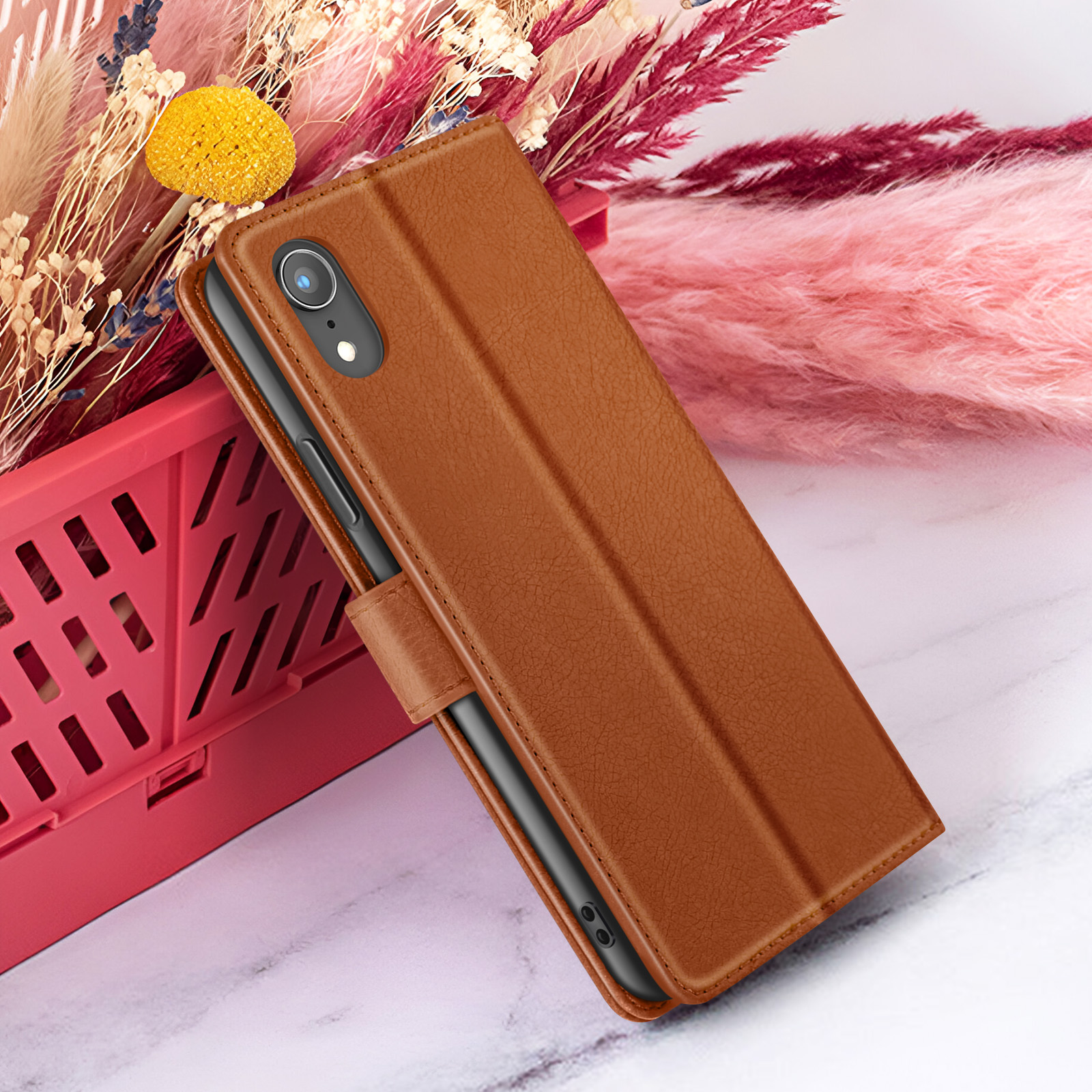 Un étui de téléphone marron sur une surface en marbre. Des fleurs séchées roses et rouges et un panier rouge en arrière-plan.