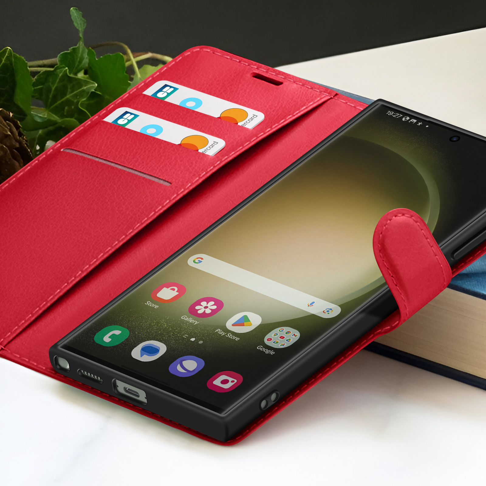 Een smartphone in een rode portemonnee, met zichtbare kaarten. Telefoon toont app-iconen en tijd. Groene bladeren en boeken op de achtergrond.