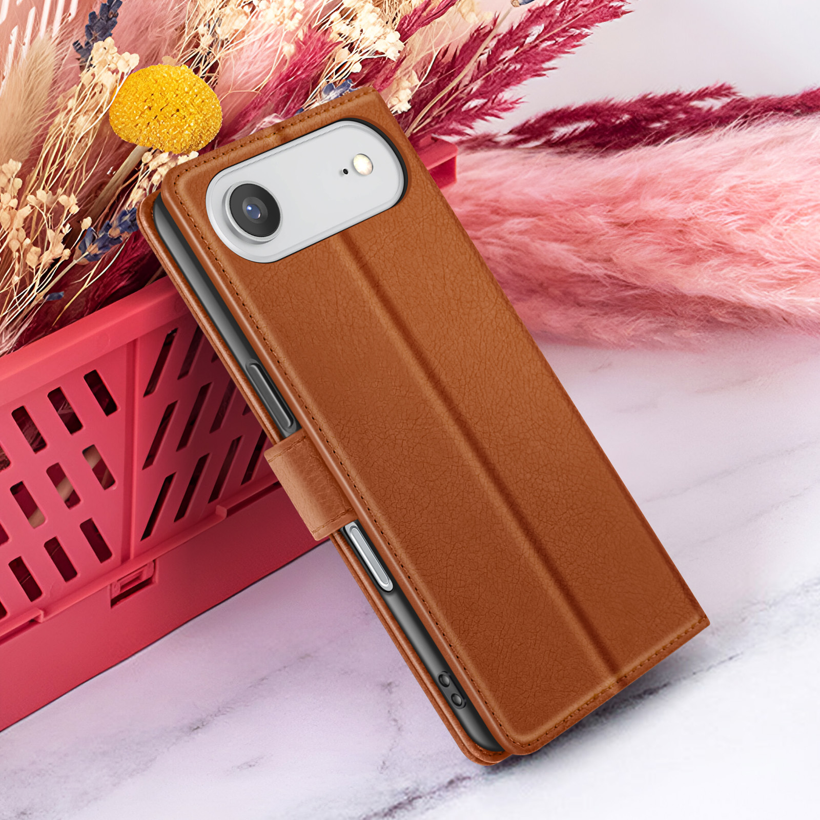 Un étui de téléphone en cuir marron est appuyé contre un panier rouge. Des fleurs séchées roses, beiges et jaunes sont en arrière-plan.