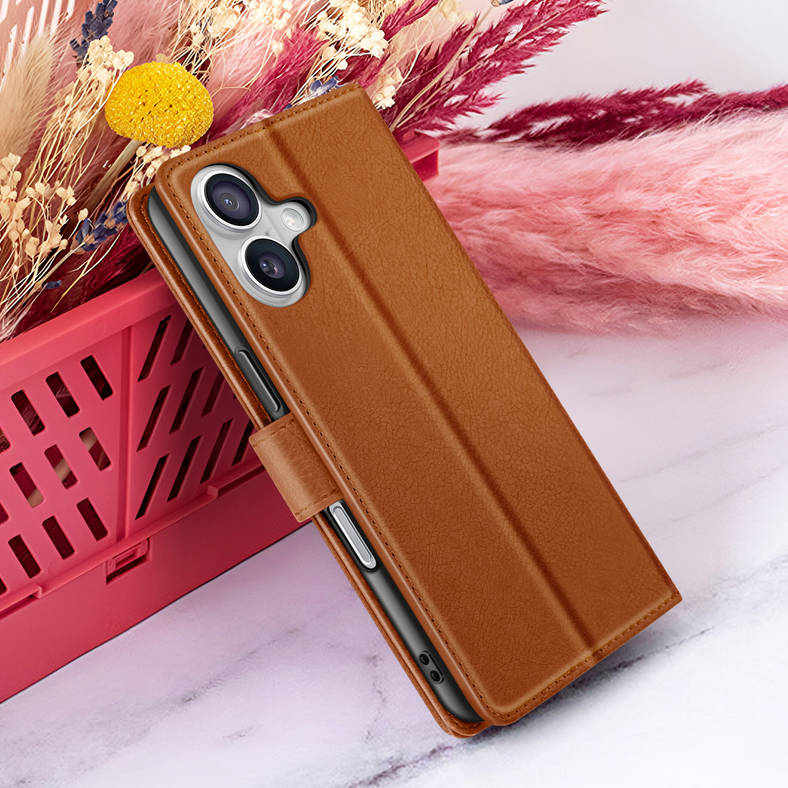 Un étui de téléphone marron est sur une caisse rouge avec des fleurs séchées en arrière-plan. Le téléphone a deux caméras.