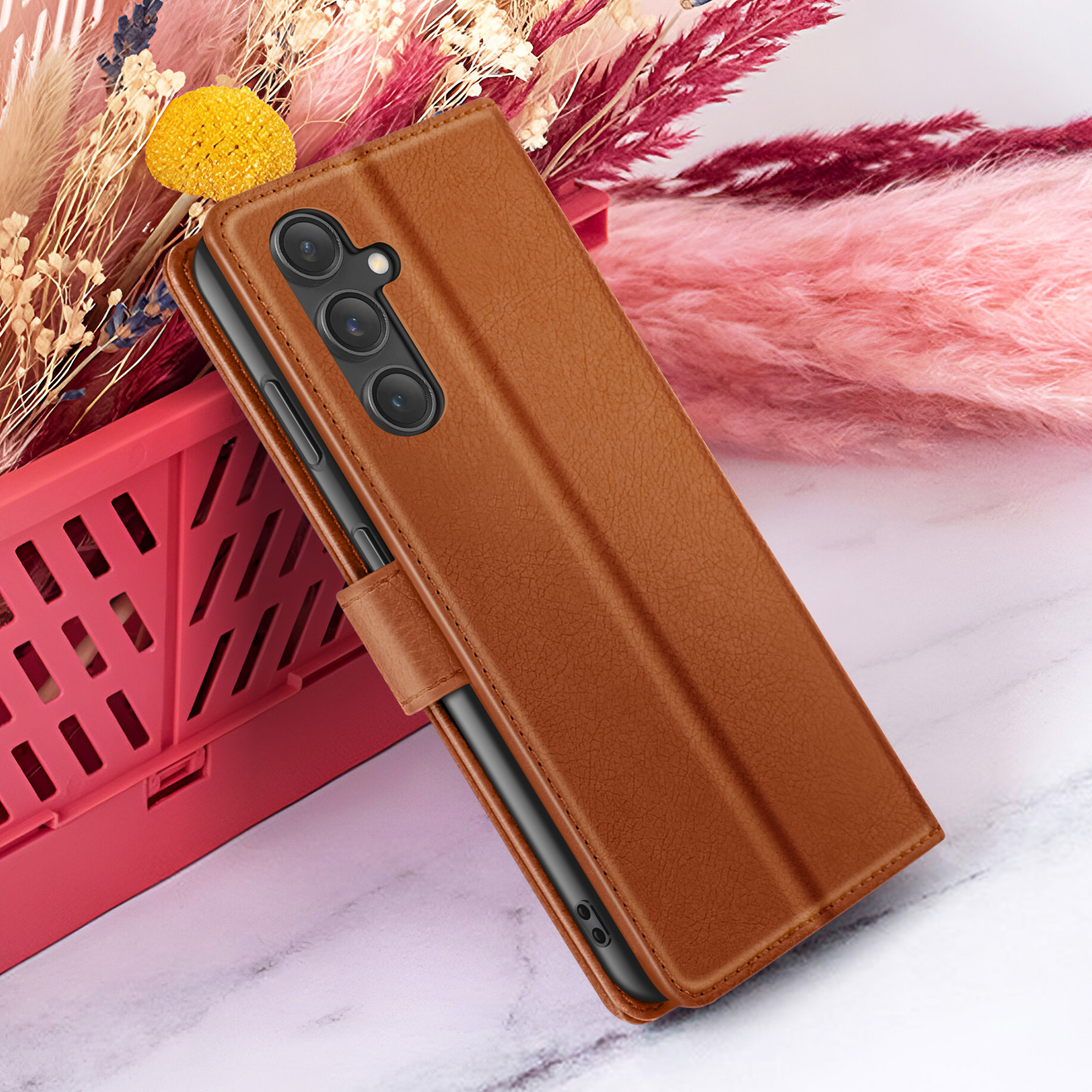 Bruine telefoonhoes op roze bloemen en een rode container, marmeren achtergrond. De hoes heeft een cameramodule.