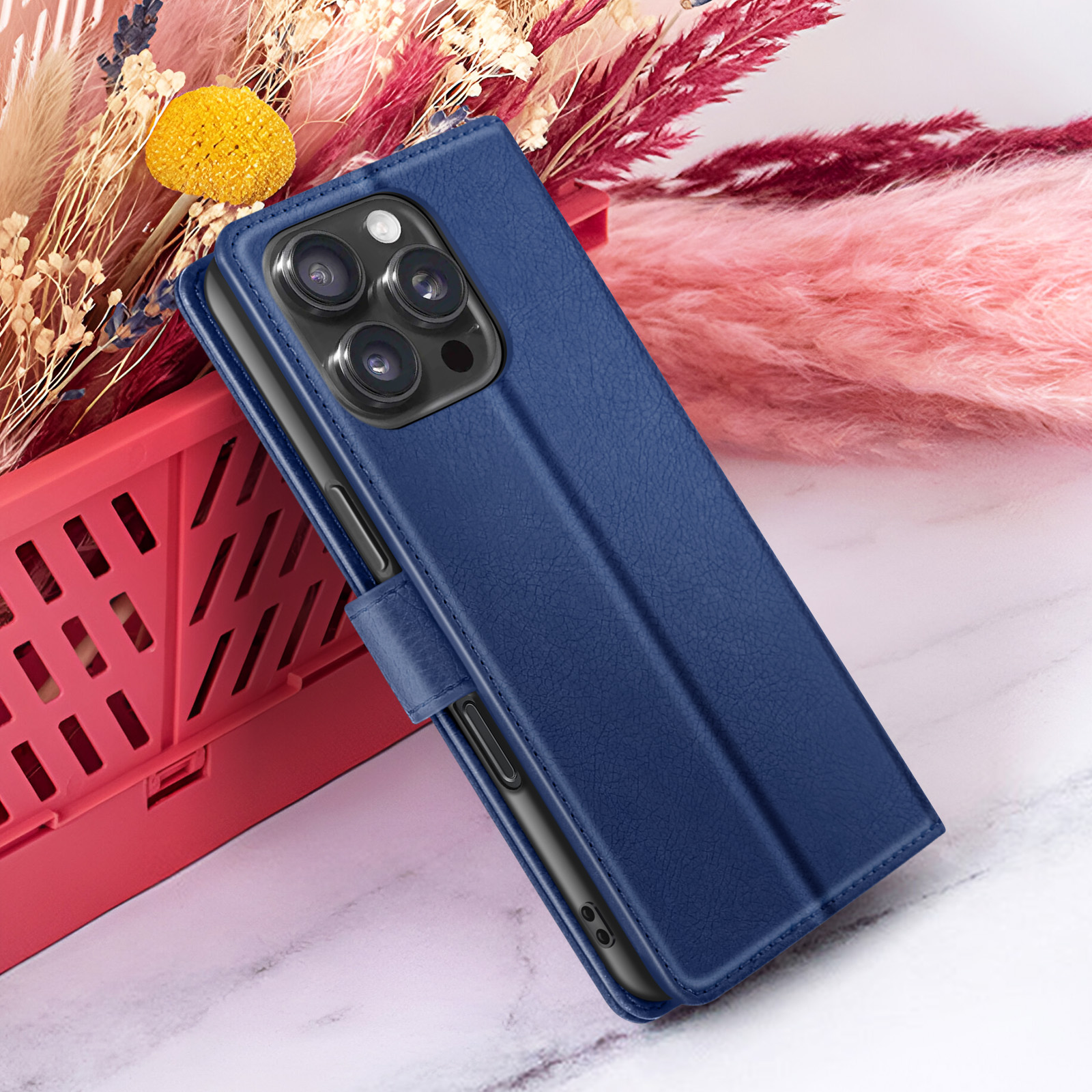 Un étui de téléphone bleu est ouvert, montrant un téléphone avec trois caméras. Le fond comprend des fleurs roses et beiges et un panier rouge.