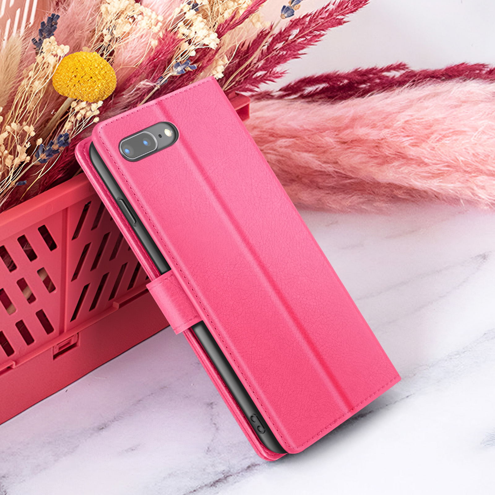 Una custodia rosa con un telefono dentro, posta davanti a fiori rosa e un cestino rosso su una superficie di marmo.