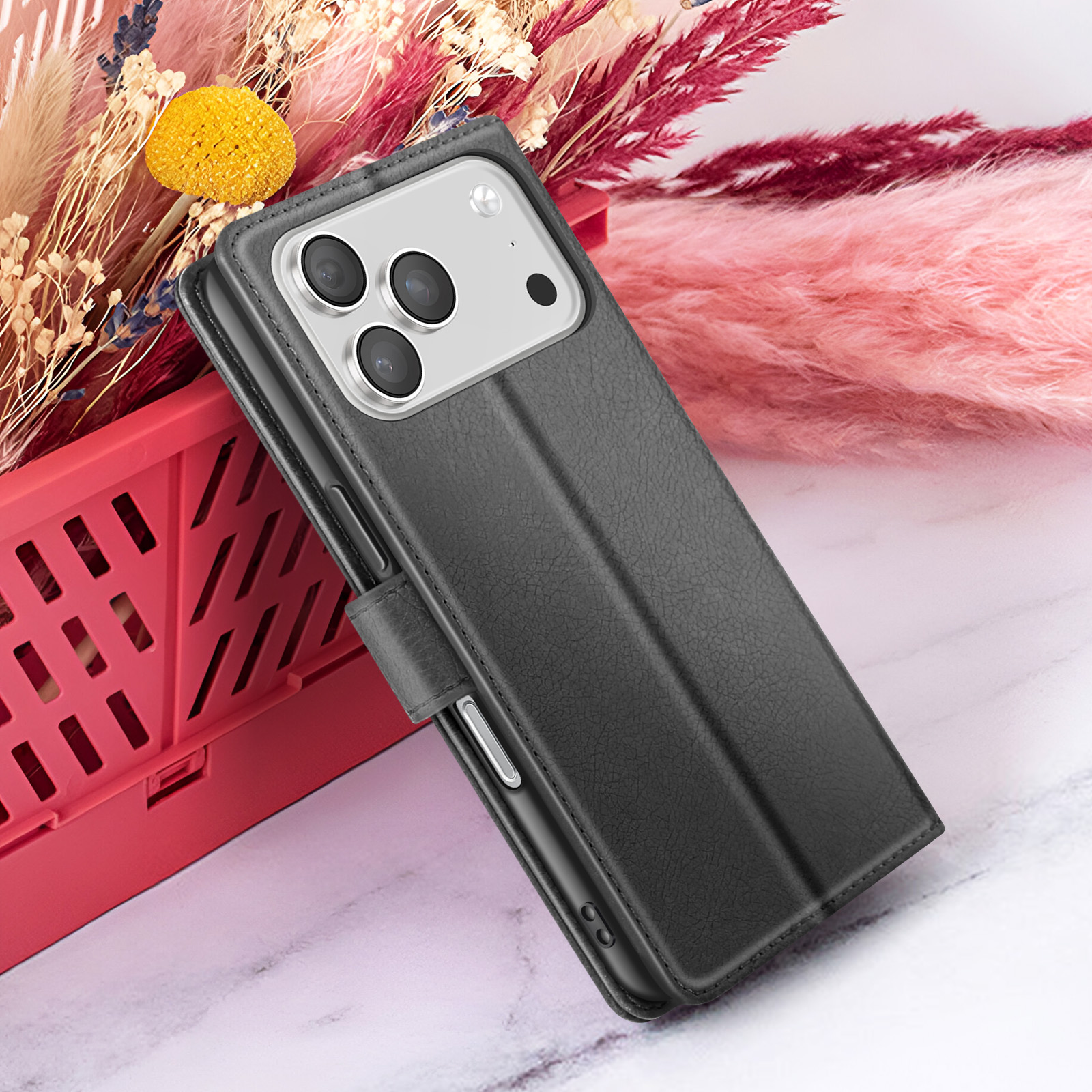 Een zwarte telefoonhoes met een zilveren cameragedeelte ligt op een rode container. Gedroogde bloemen in roze en beige staan op de achtergrond.