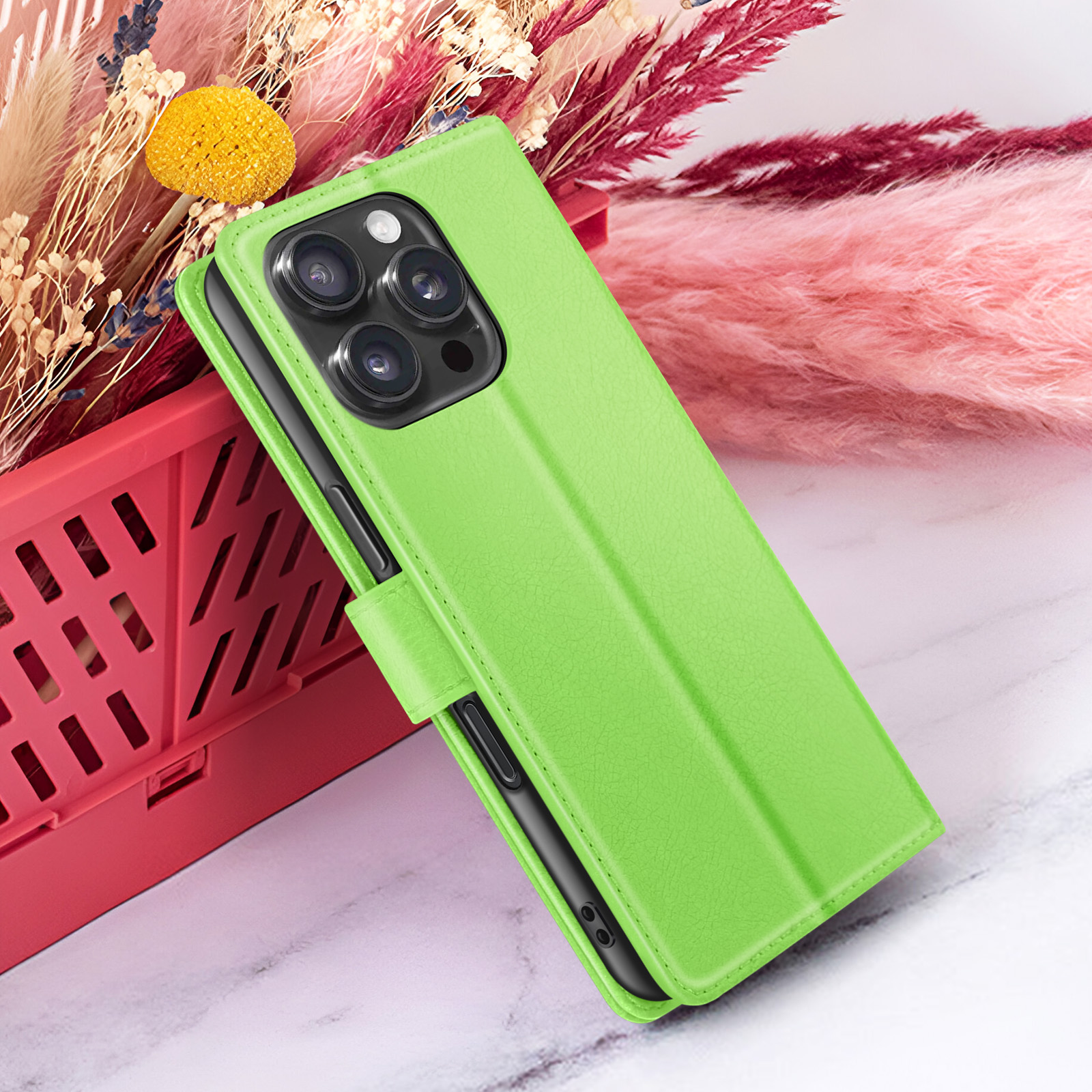 Groene telefoonhoes met drie cameralenzen. Roze en bordeauxrode bloemen op de achtergrond. De hoes zit in een rode mand.