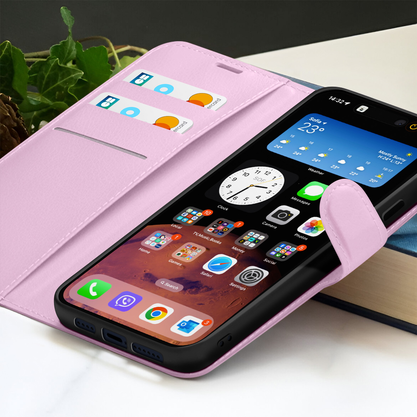 Een iPhone in een roze hoesje toont apps en weer. Creditcards zijn zichtbaar in de sleuven van de hoes. Een boek staat op de achtergrond.
