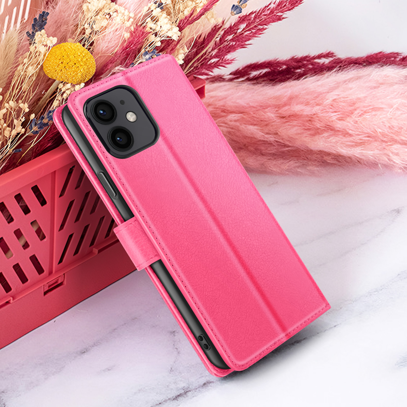 Custodia rosa per telefono con telefono dentro, su superficie in marmo bianco, fiori secchi e cestino rosa sullo sfondo.