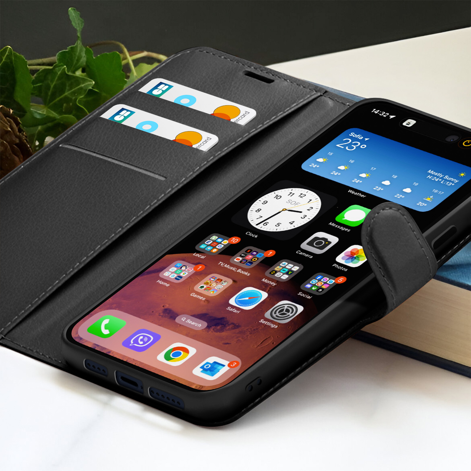 Un telefono nero in una custodia nera mostra le app. Due carte sono nella custodia. Sfondo: una pianta, un libro.