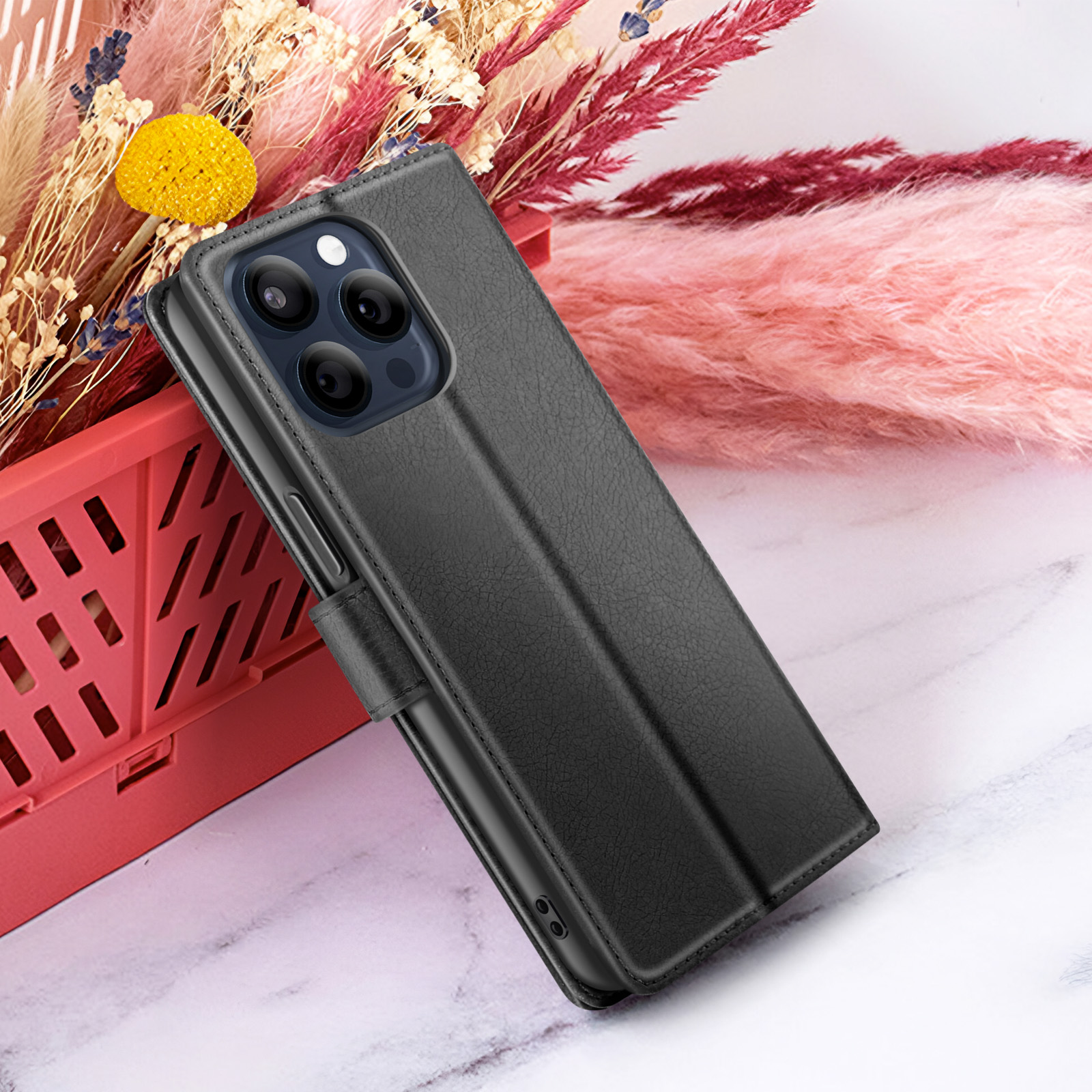 Custodia nera per telefono con più fotocamere, appoggiata a fiori in un cesto rosso e su una superficie di marmo bianco.