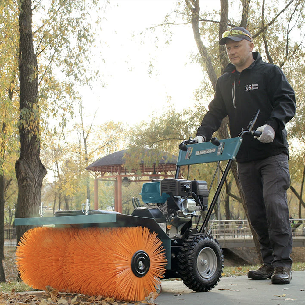 Un homme utilise une balayeuse avec une brosse orange. La machine a un cadre bleu-vert. Des feuilles d'automne sont au sol. Arbres et un gazebo en arrière-plan.