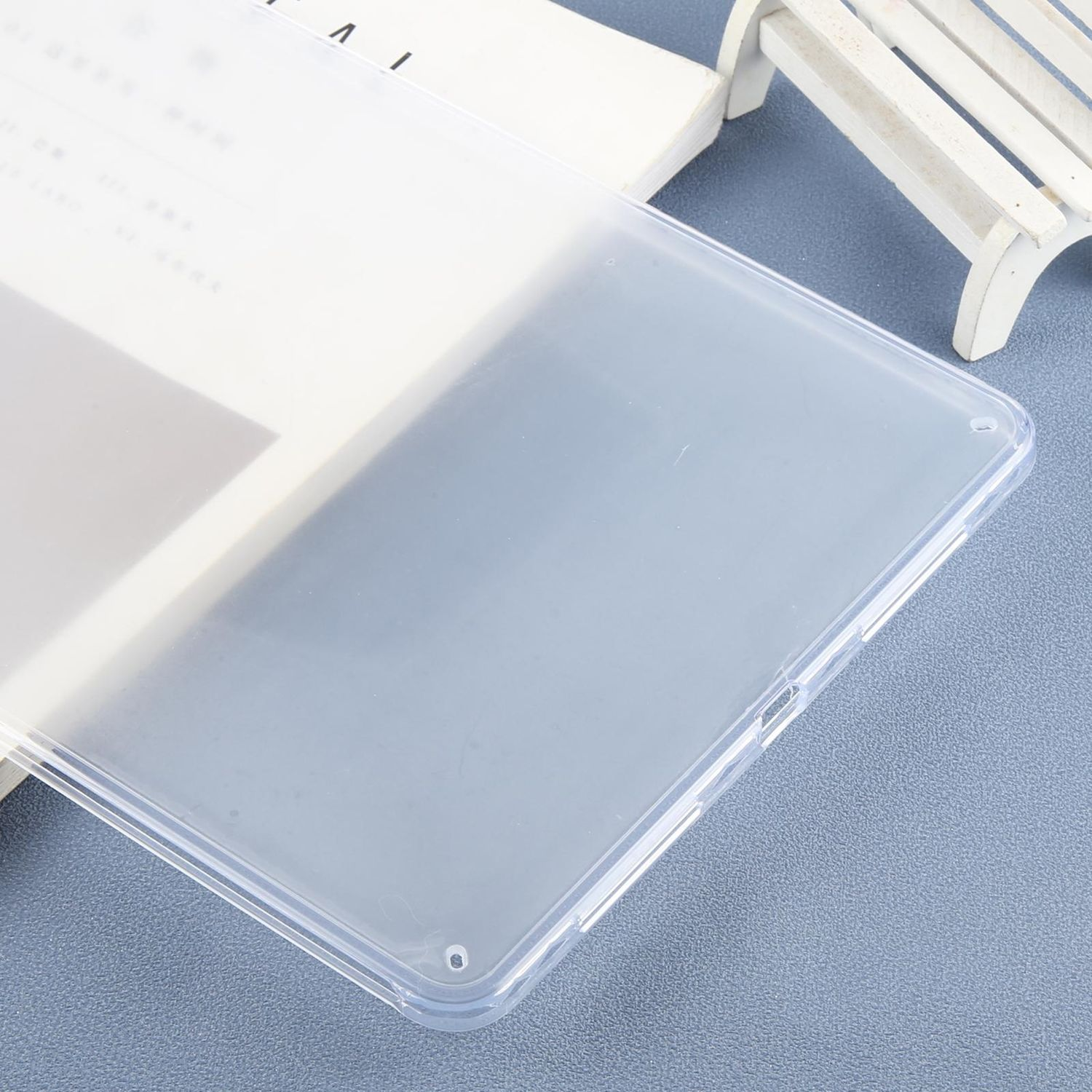 Eine transparente Tablet-Hülle liegt auf einem Tisch mit einem Buch und einer kleinen weißen Bank im Hintergrund, alles auf einer blauen Oberfläche.