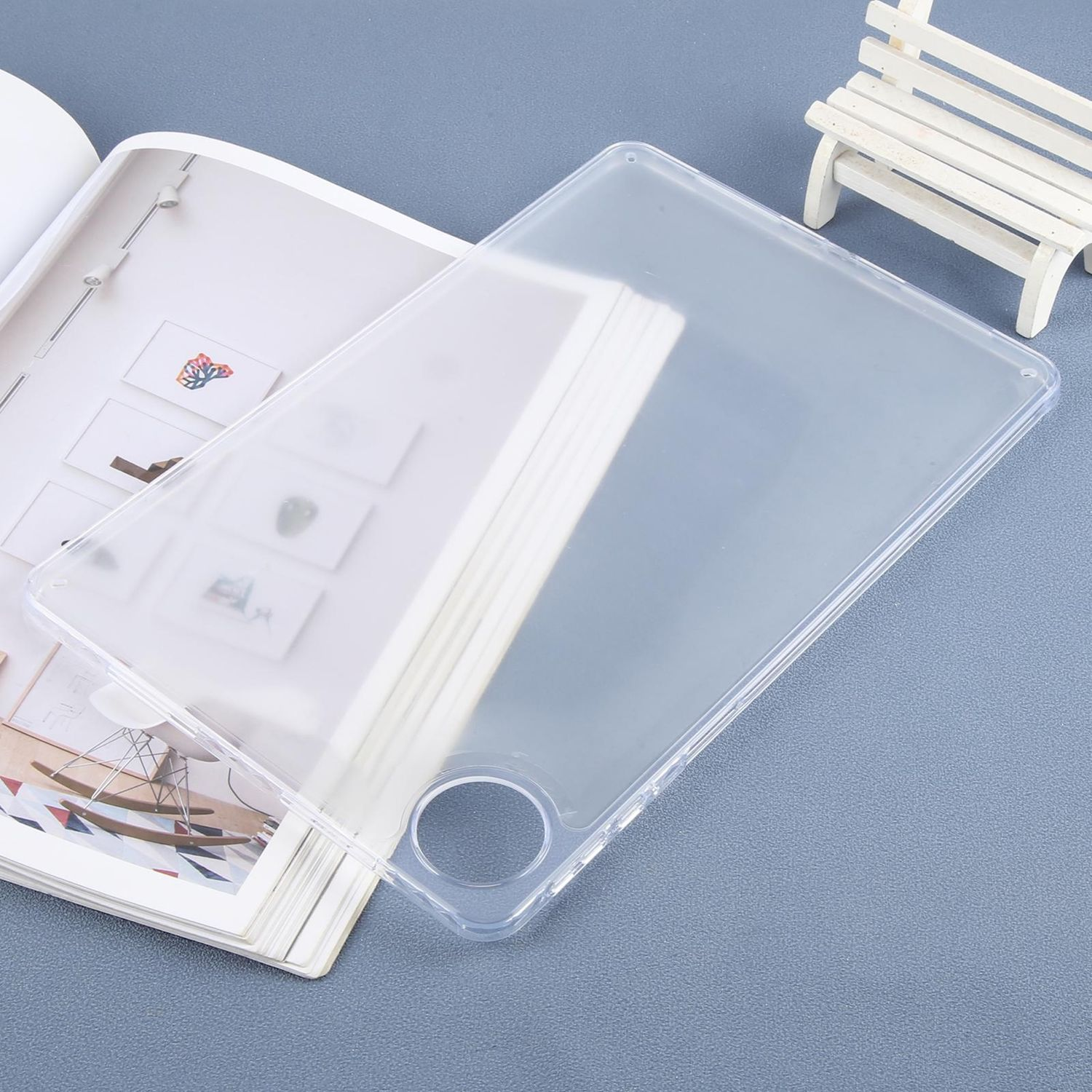 Eine transparente Tablethülle liegt auf einem offenen Magazin. Eine kleine weiße Bank steht im Hintergrund auf einer blauen Oberfläche.