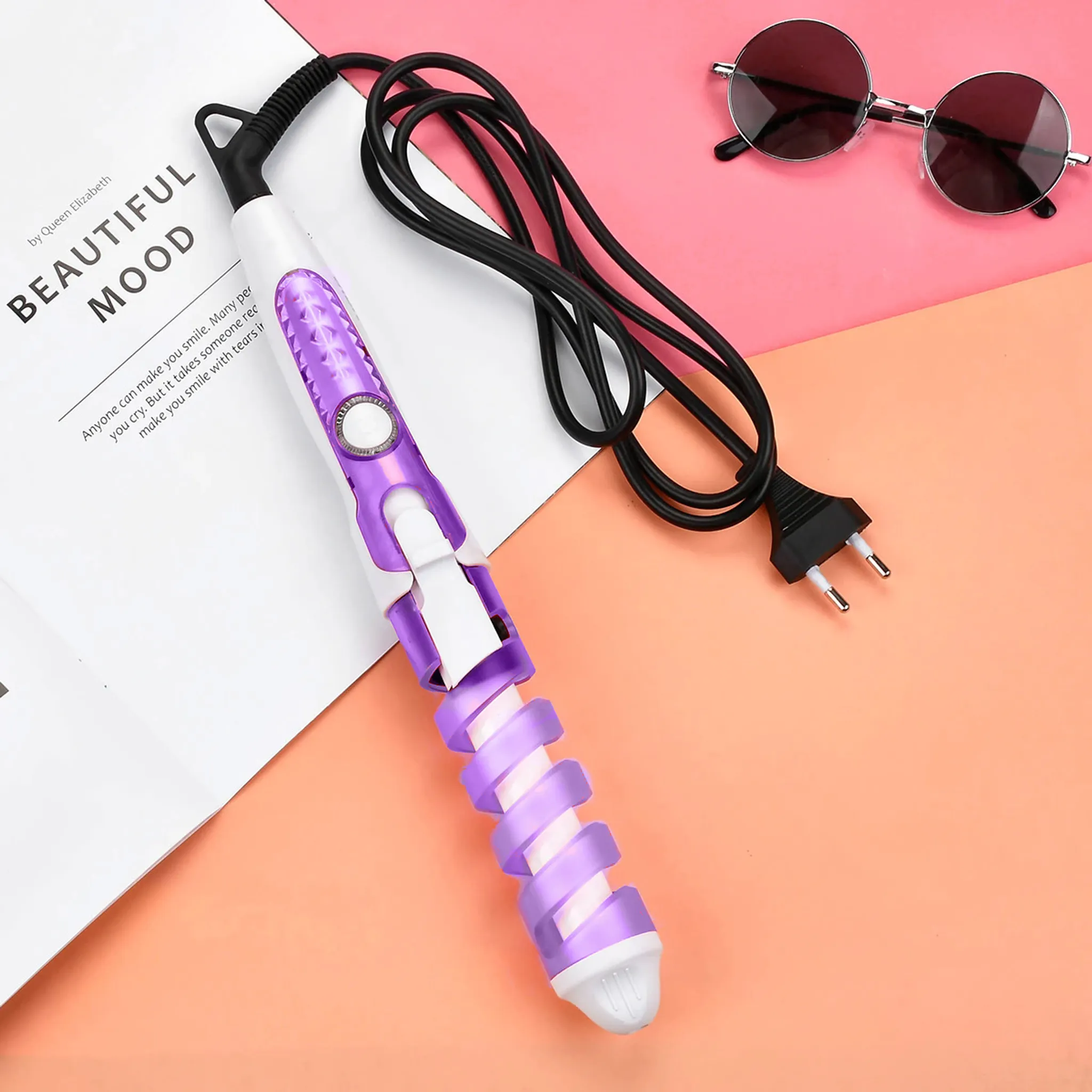 Ein Lockenstab, eine Sonnenbrille und ein Netzkabel liegen auf einer rosa-pfirsichfarbenen Oberfläche. Text auf weißem Papier.