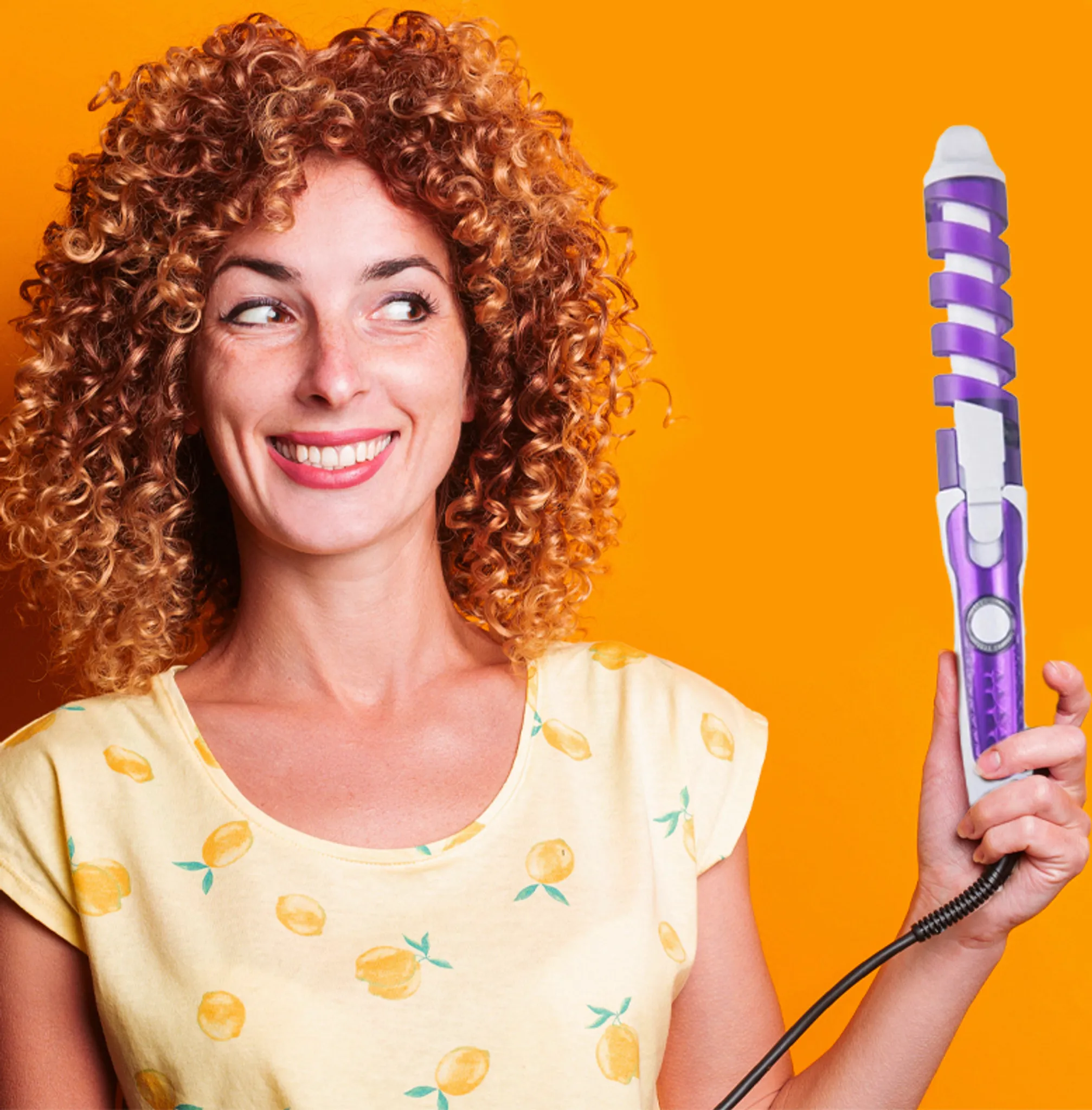Frau mit Lockenstab lächelt. Gelbes Shirt mit Zitronen, orangefarbener Hintergrund. Lockiges Haar. Eisen ist weiß und lila.