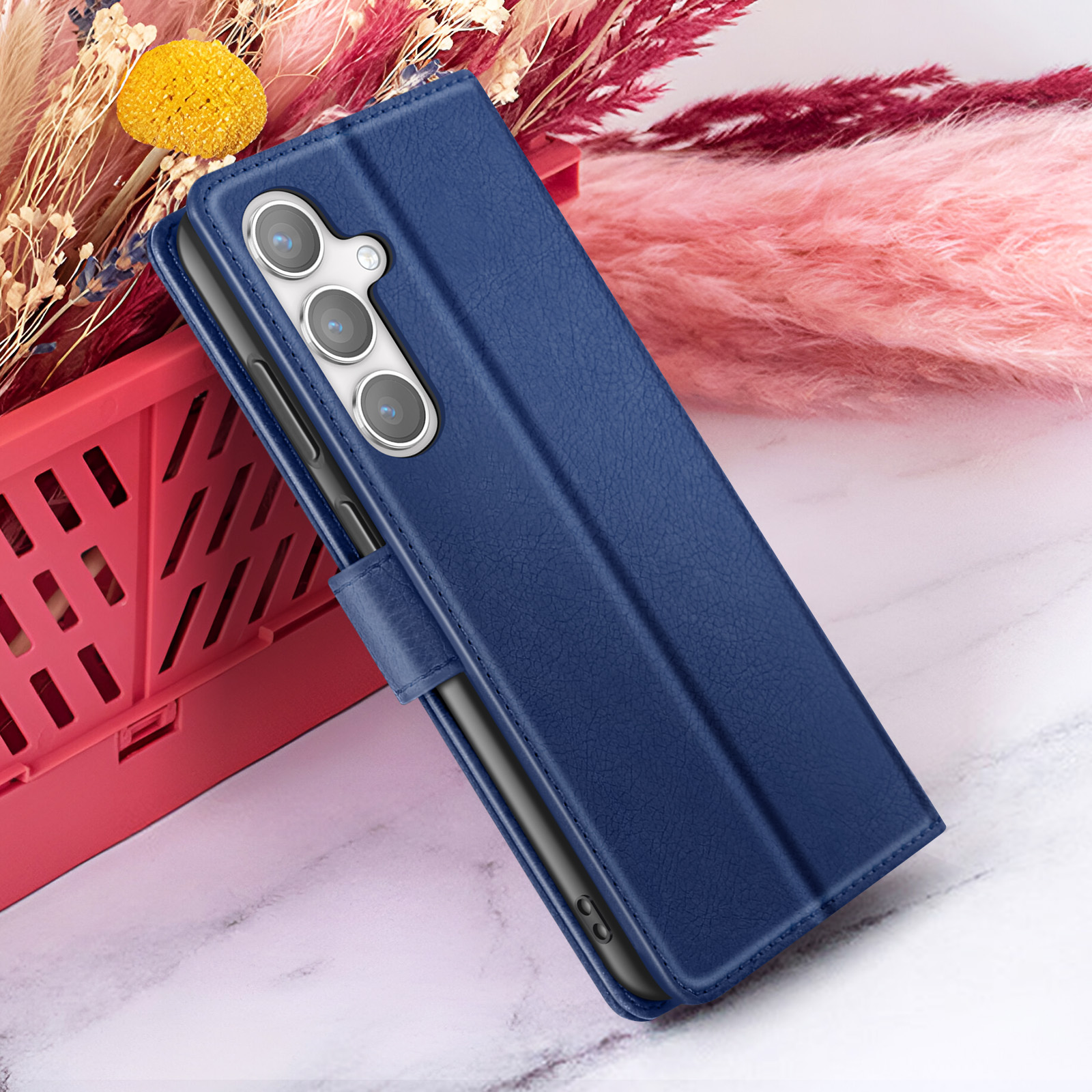 Een blauwe telefoonhoes met een zwarte telefoon erin. Roze en rode gedroogde bloemen en een rode mand op de achtergrond.