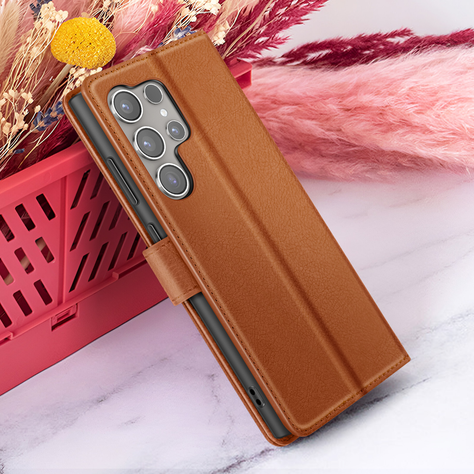 Bruine leren telefoonhoes met meerdere camera's, naast bloemen in een rode mand op marmer.