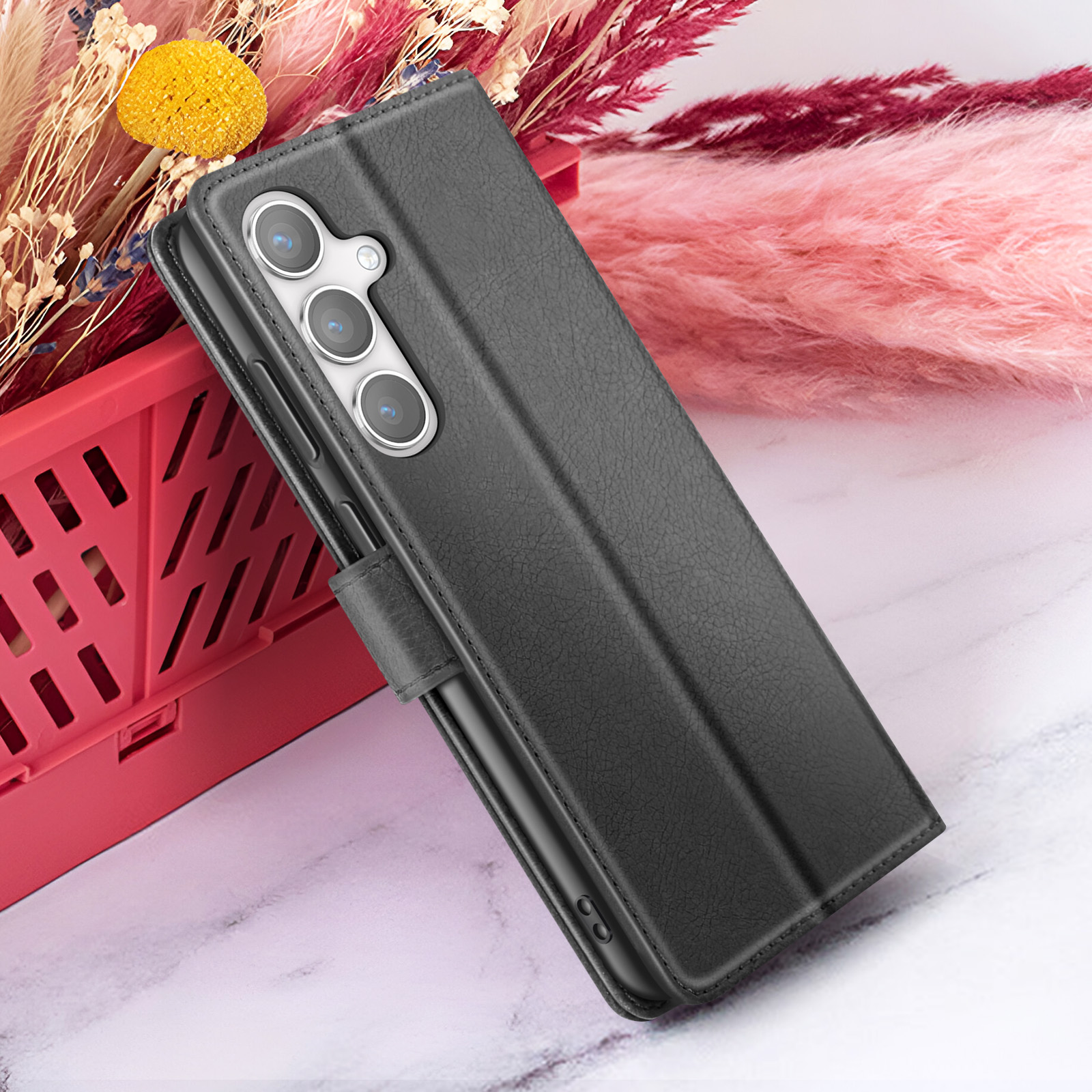 Een zwarte telefoonhoes leunt op een rode container. Roze bloemen op de achtergrond. De hoes heeft drie camera's.