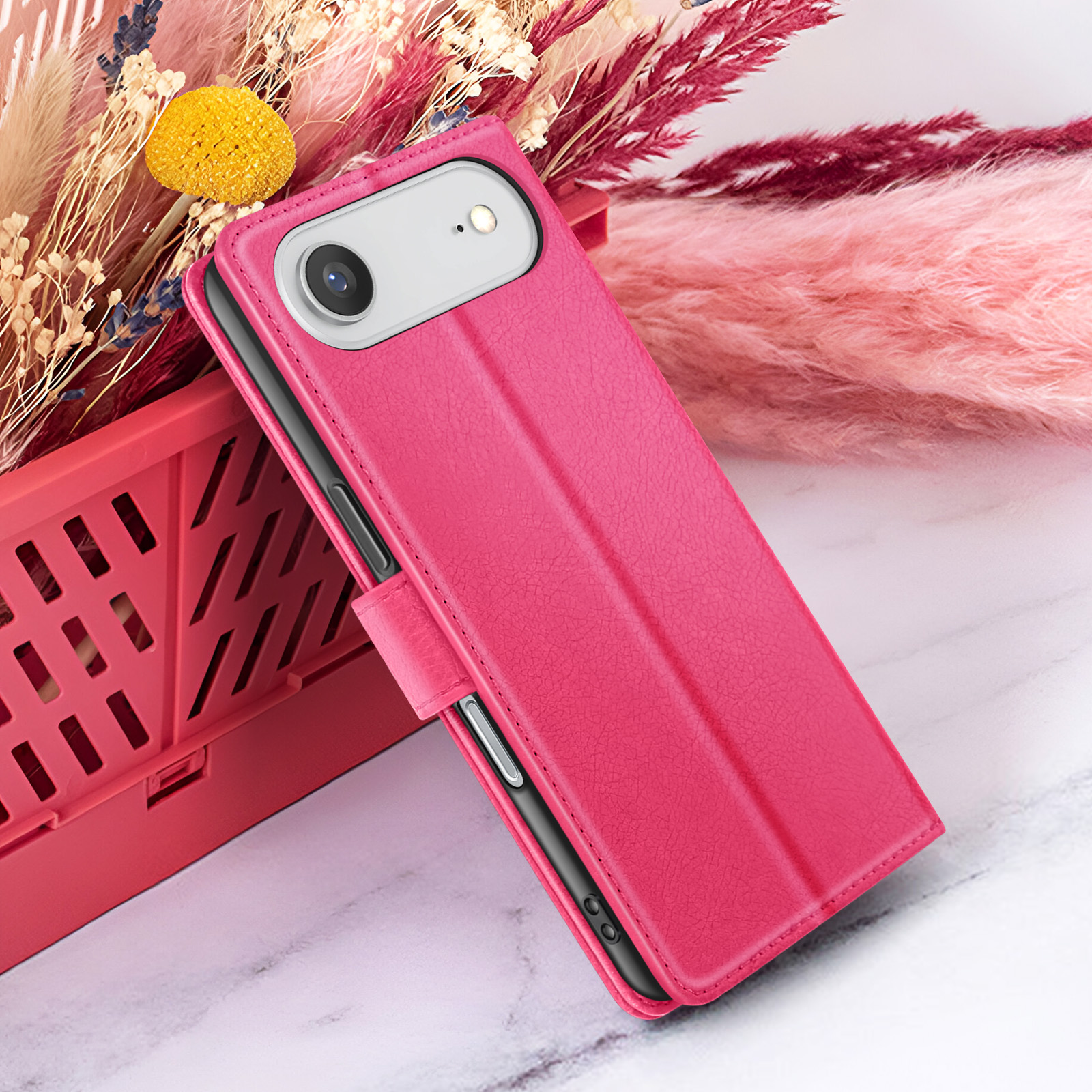 Un étui de téléphone rose avec une caméra est près de fleurs séchées dans une boîte rouge. Le fond a des teintes roses et violettes.