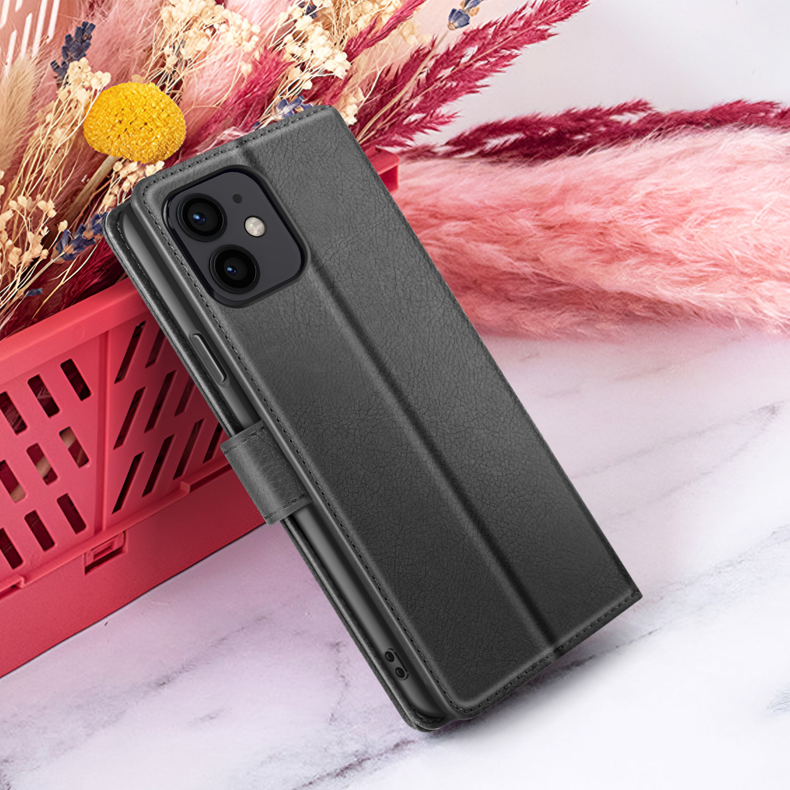 Una custodia nera per telefono è esposta, parzialmente aperta. Lo sfondo include fiori rosa, un cestino rosso e una superficie marmorea.
