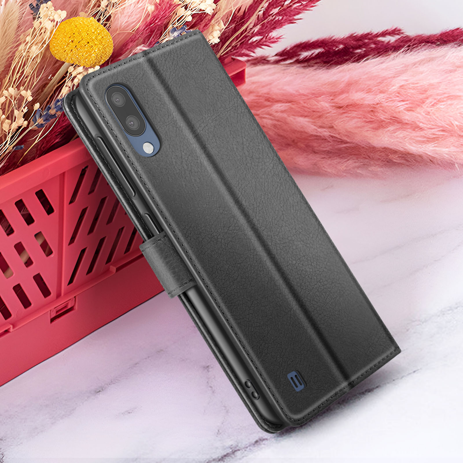 Étui de téléphone noir sur surface en marbre, avec panier rouge et fleurs roses et blanches en arrière-plan.