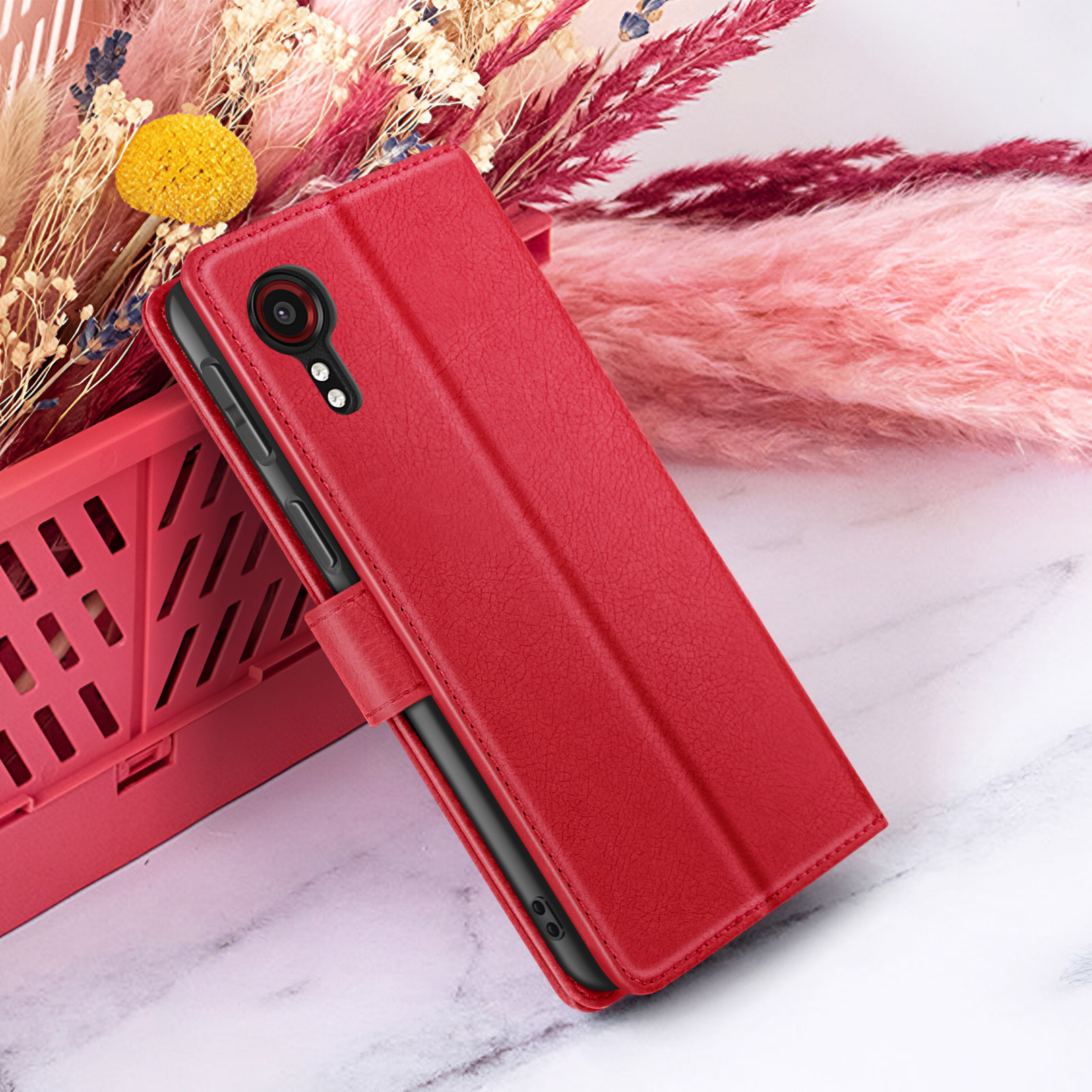Een rode telefoonhoes wordt getoond, met een telefoon erin. Gedroogde bloemen en een roze achtergrond.