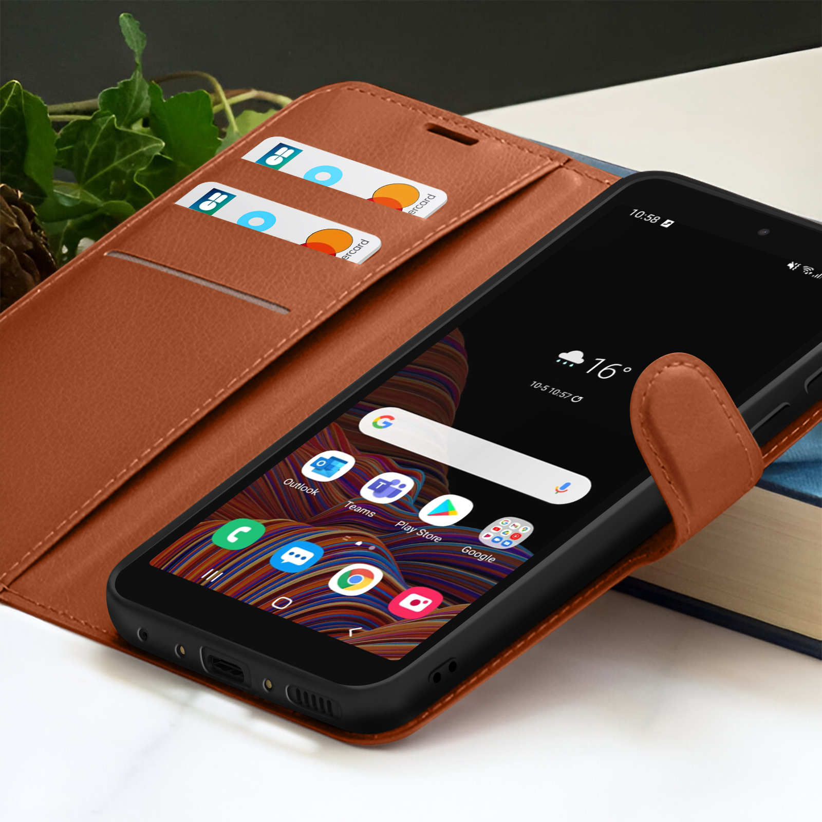 Een smartphone in een bruine leren hoes toont apps en het weer. Creditcards in de sleuven van de hoes, met een boek op de achtergrond.