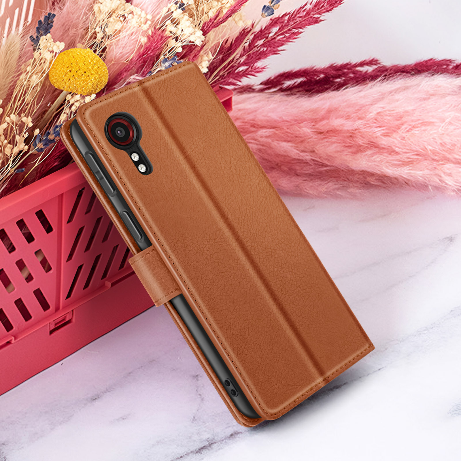 Een bruine telefoonhoes met een telefoon erin ligt op een marmeren oppervlak. Bloemen op de achtergrond.
