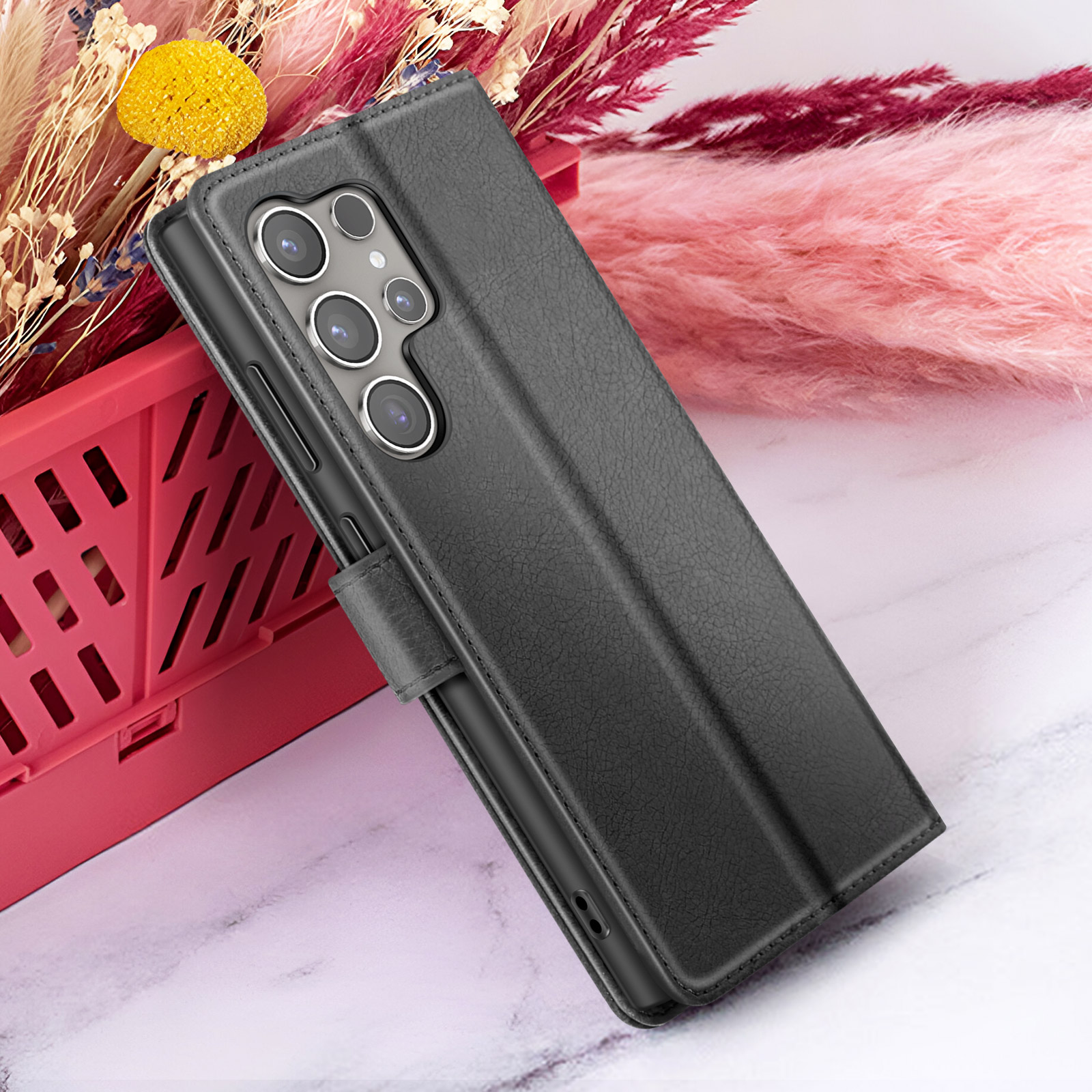 Zwarte telefoonhoes met cameralenzen, leunend op een rode mand met bloemen op de achtergrond op een marmeren oppervlak.